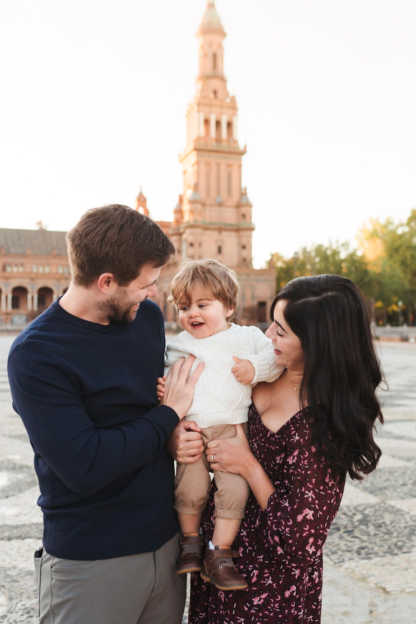 A young family with a toddler smiling at Plaza de España, Seville, with the iconic tower in the background.