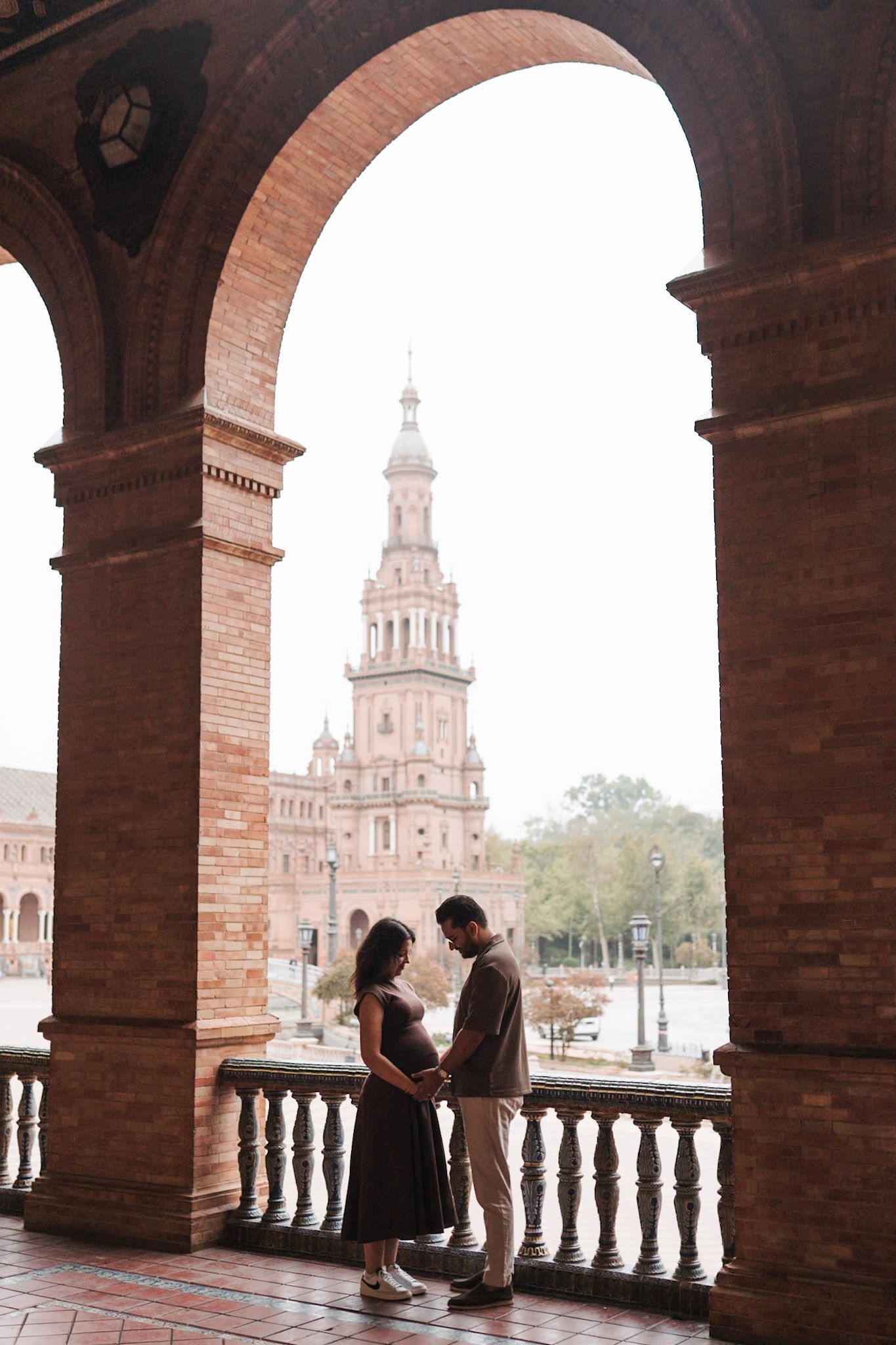 Pregnant couple embracing under brick archway at Plaza de España, Seville.