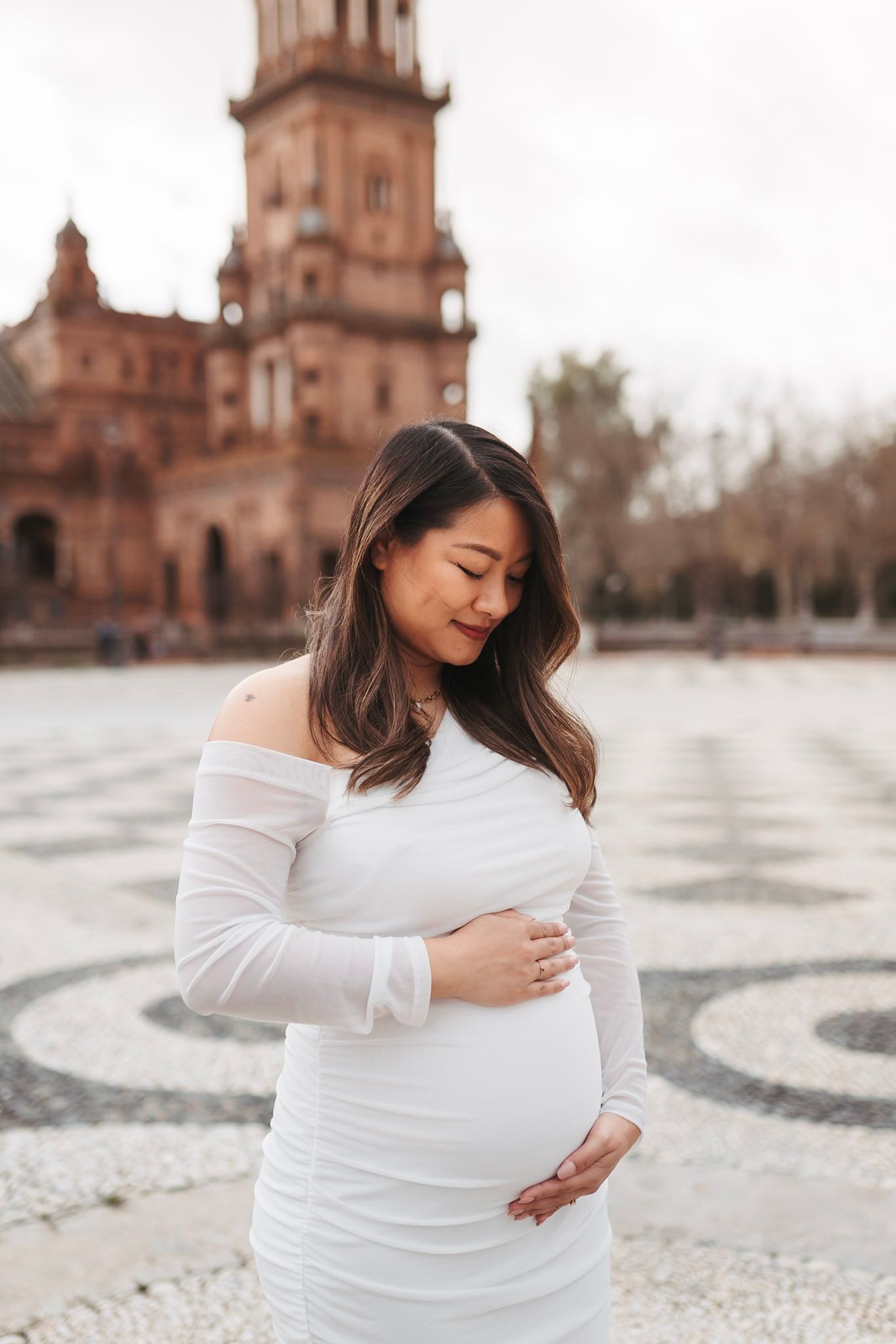 Radiant pregnant woman in white off-shoulder dress cradles baby bump before Plaza de Espana tower.