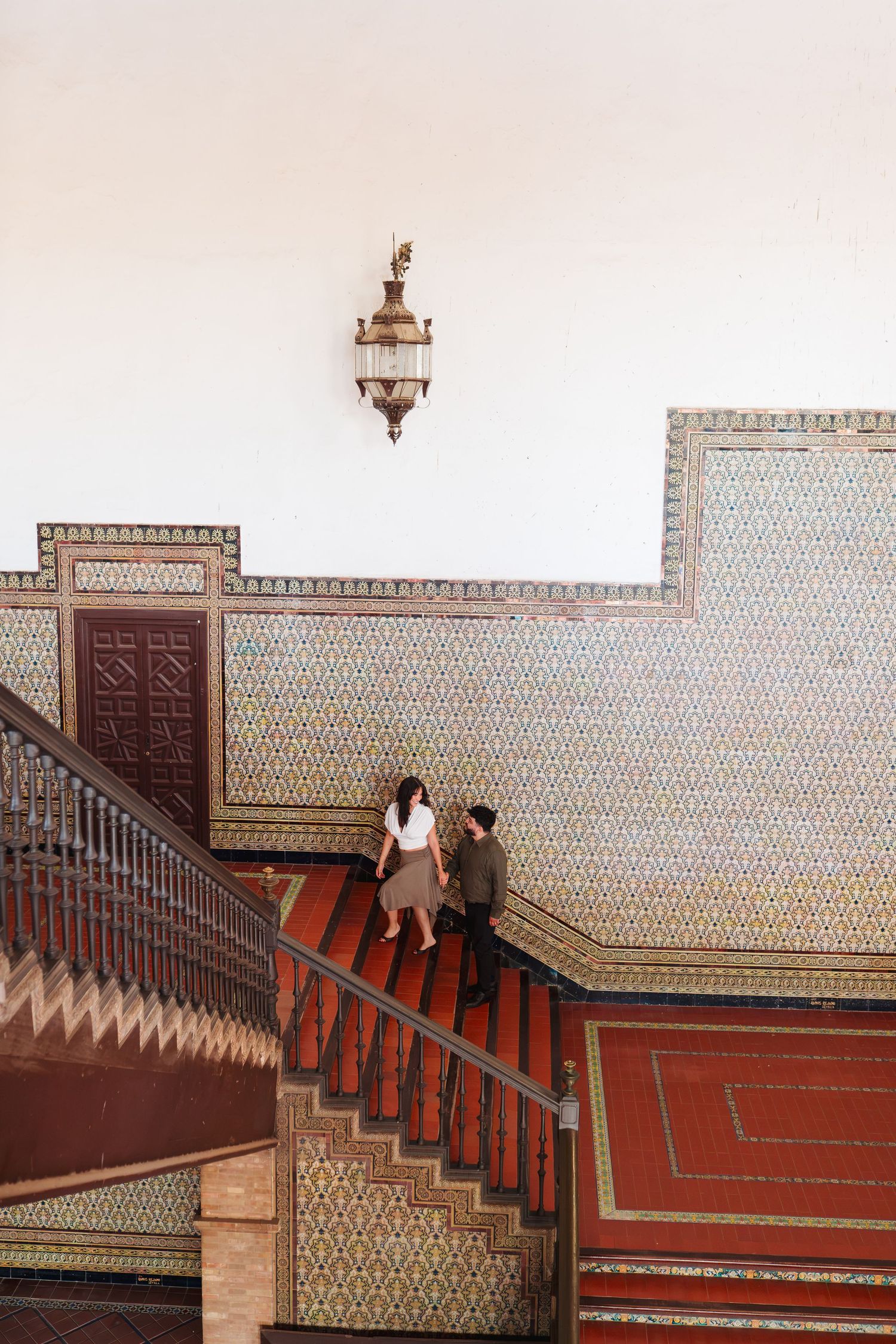 Two people descend ornate tiled staircase in a Moorish-style building with decorative geometric patterns and vintage lantern.
