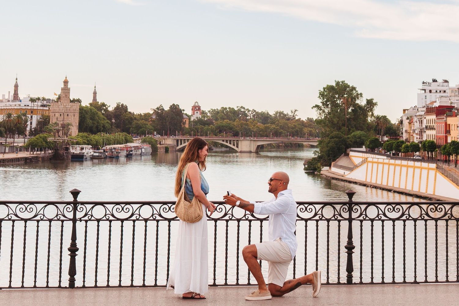 Man proposes to woman on bridge overlooking scenic river in Seville, Spain, with historic towers in background.