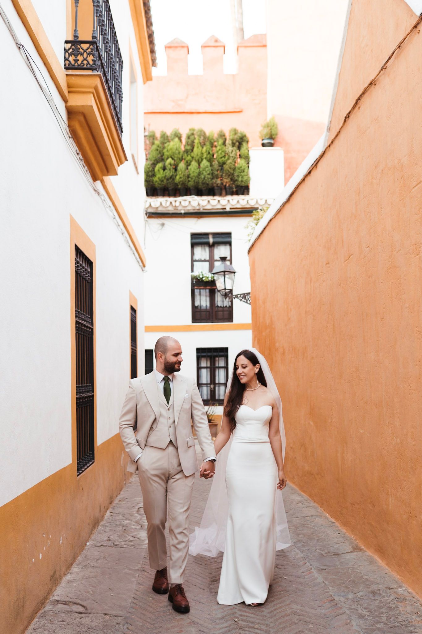 Bride and groom walking hand in hand through narrow orange and white alleyway in Seville, Spain.