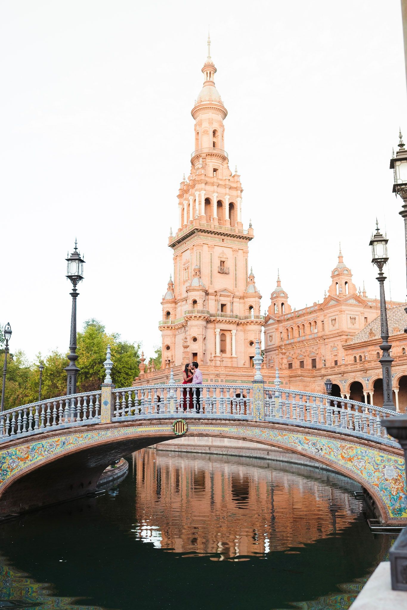 Couple standing on ornate tiled bridge at Plaza de España, Seville, with reflection in canal and baroque tower backdrop.