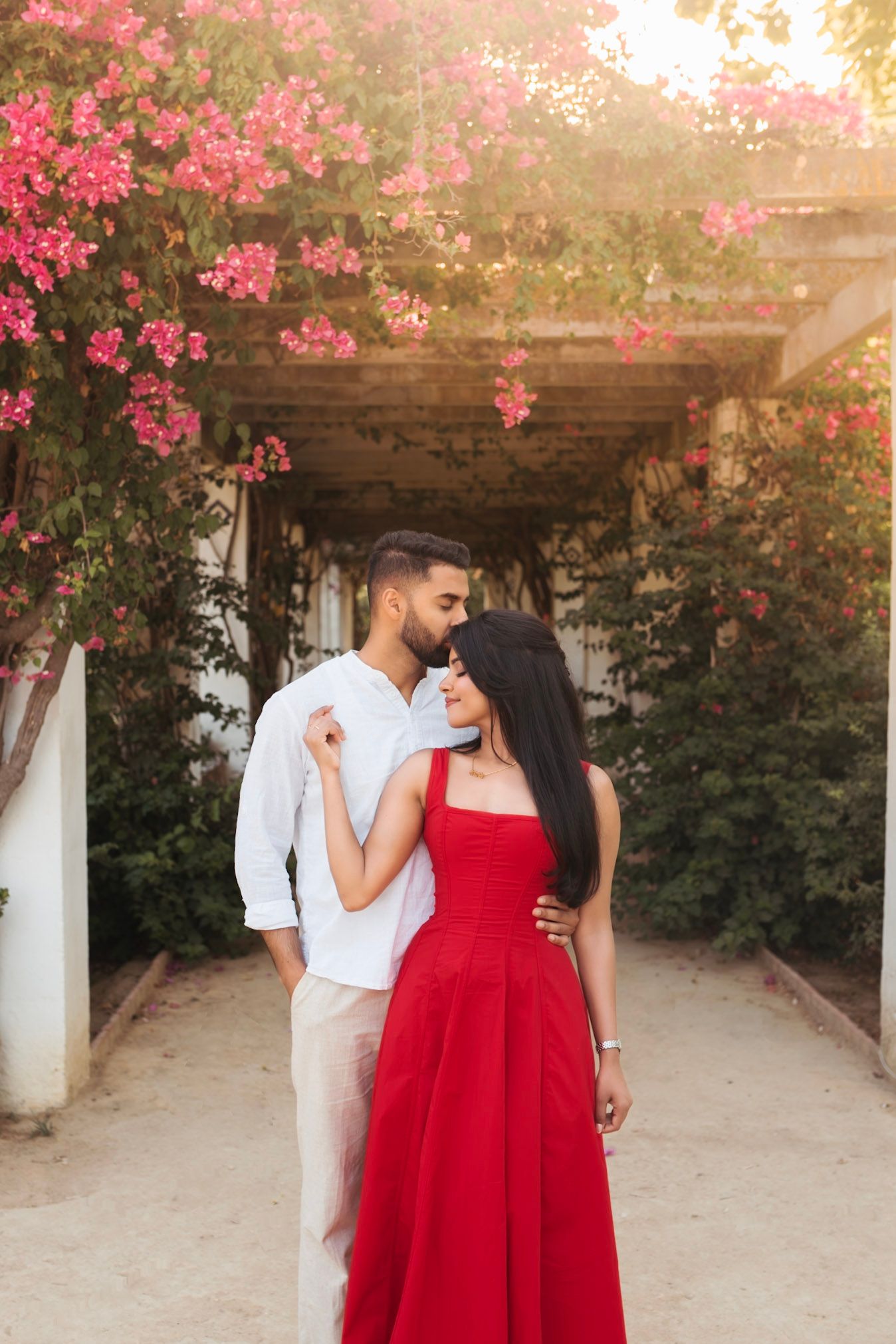 Romantic couple embracing under a pergola of vibrant pink bougainvillea flowers in warm golden sunlight.