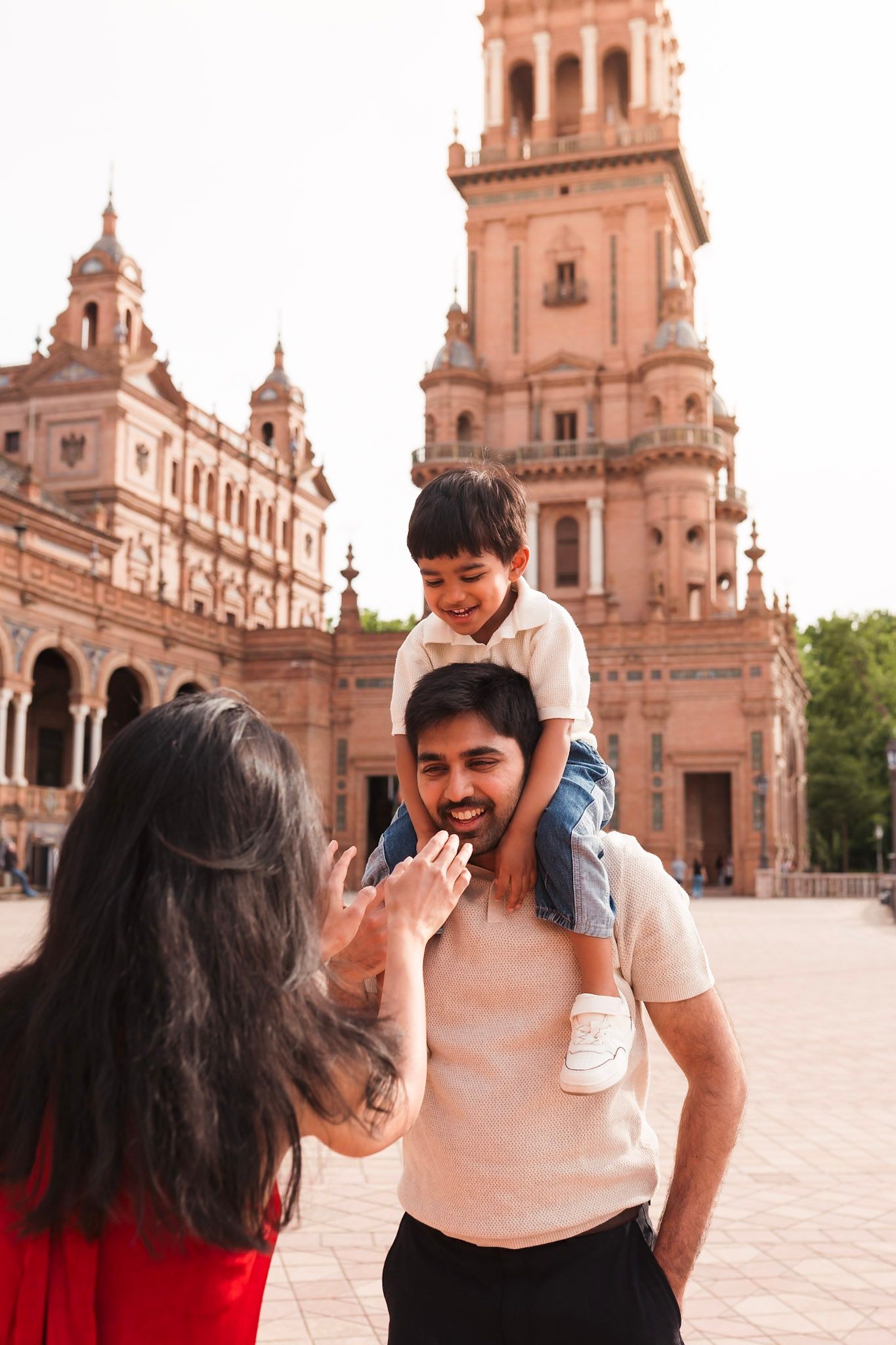 A father carries his son on his shoulders while mother smiles at Plaza de Espana in Seville.