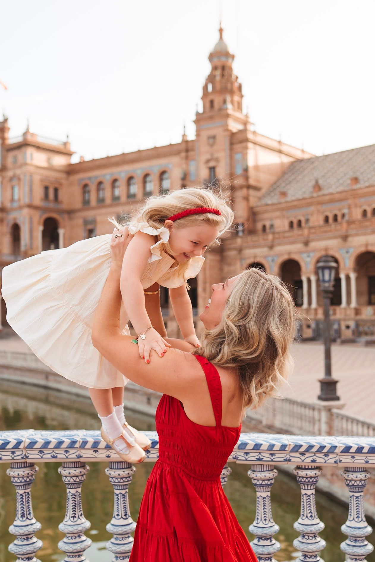 Mother in red dress lifting laughing toddler girl at Plaza de Espana in Seville, Spain.