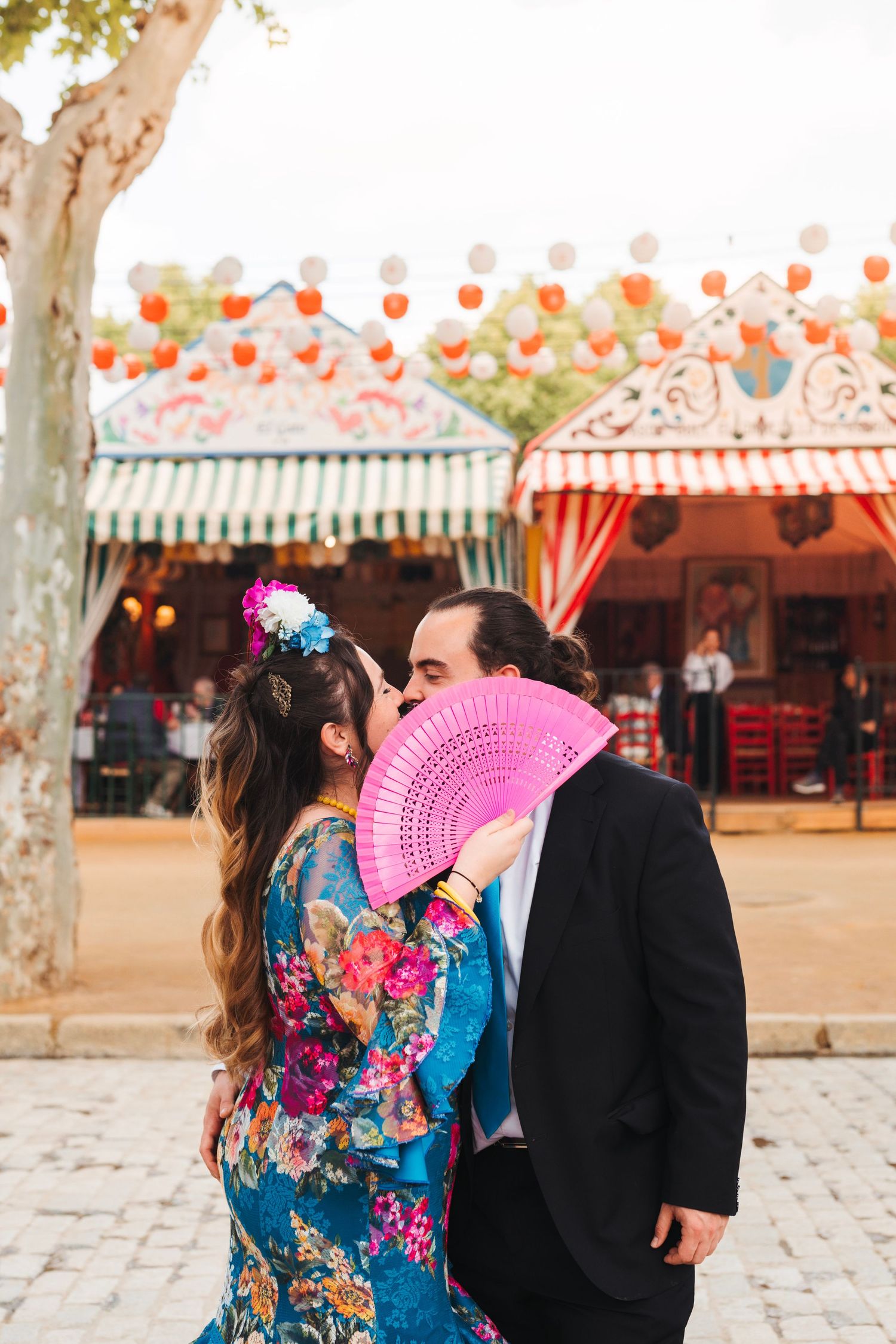 Couple kissing behind pink fan in traditional Spanish attire at colorful Feria de Abril.