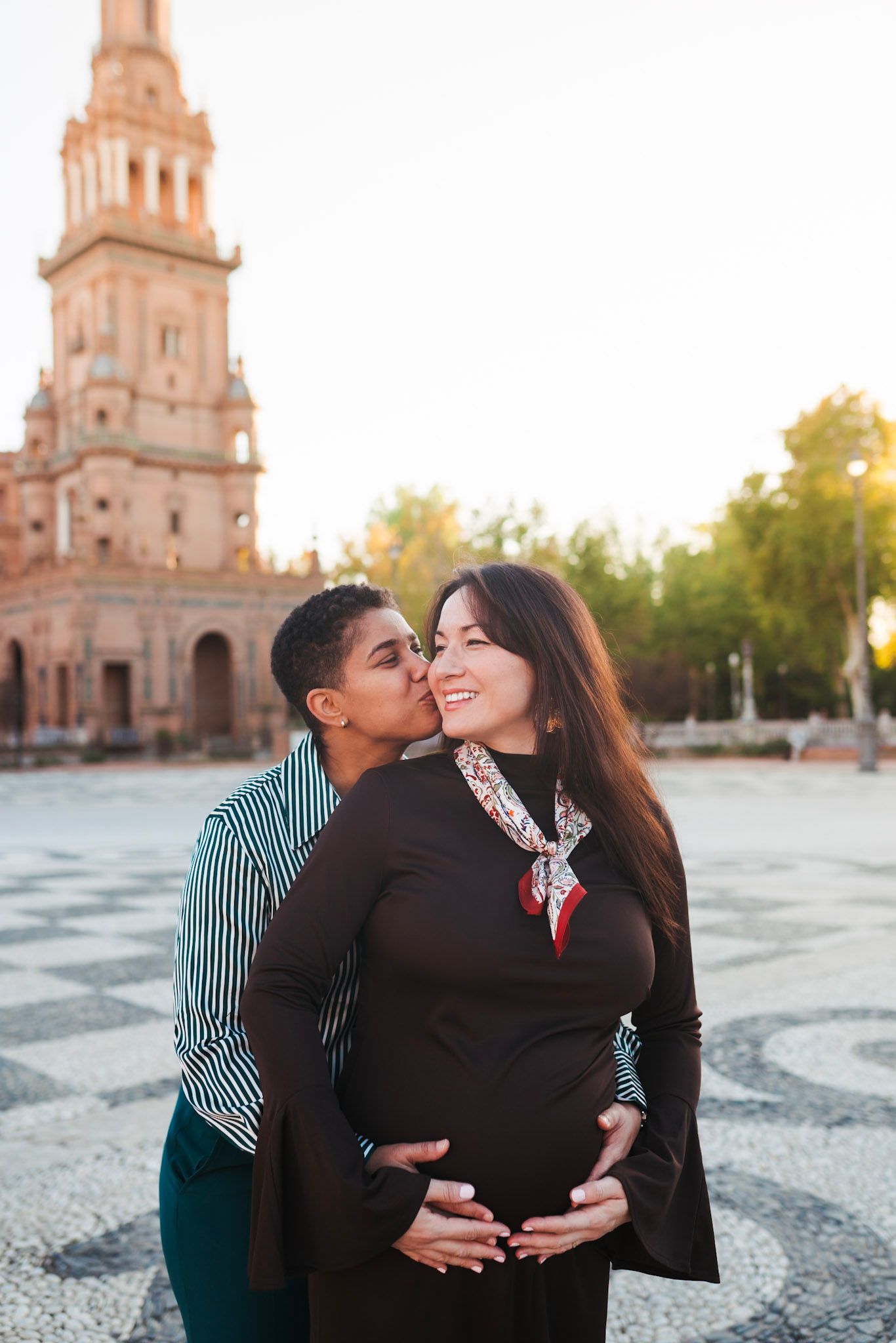 Same-sex couple maternity photo with kiss on cheek at Plaza de España Seville.