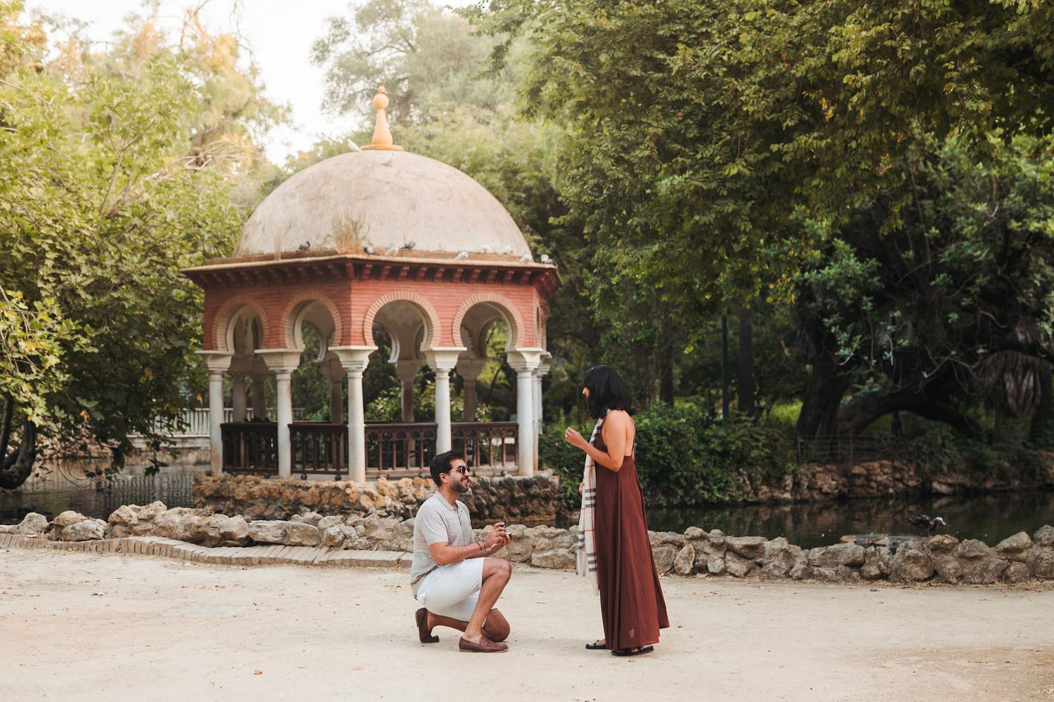 Man proposes on one knee to woman in burgundy dress near ornate gazebo in park.