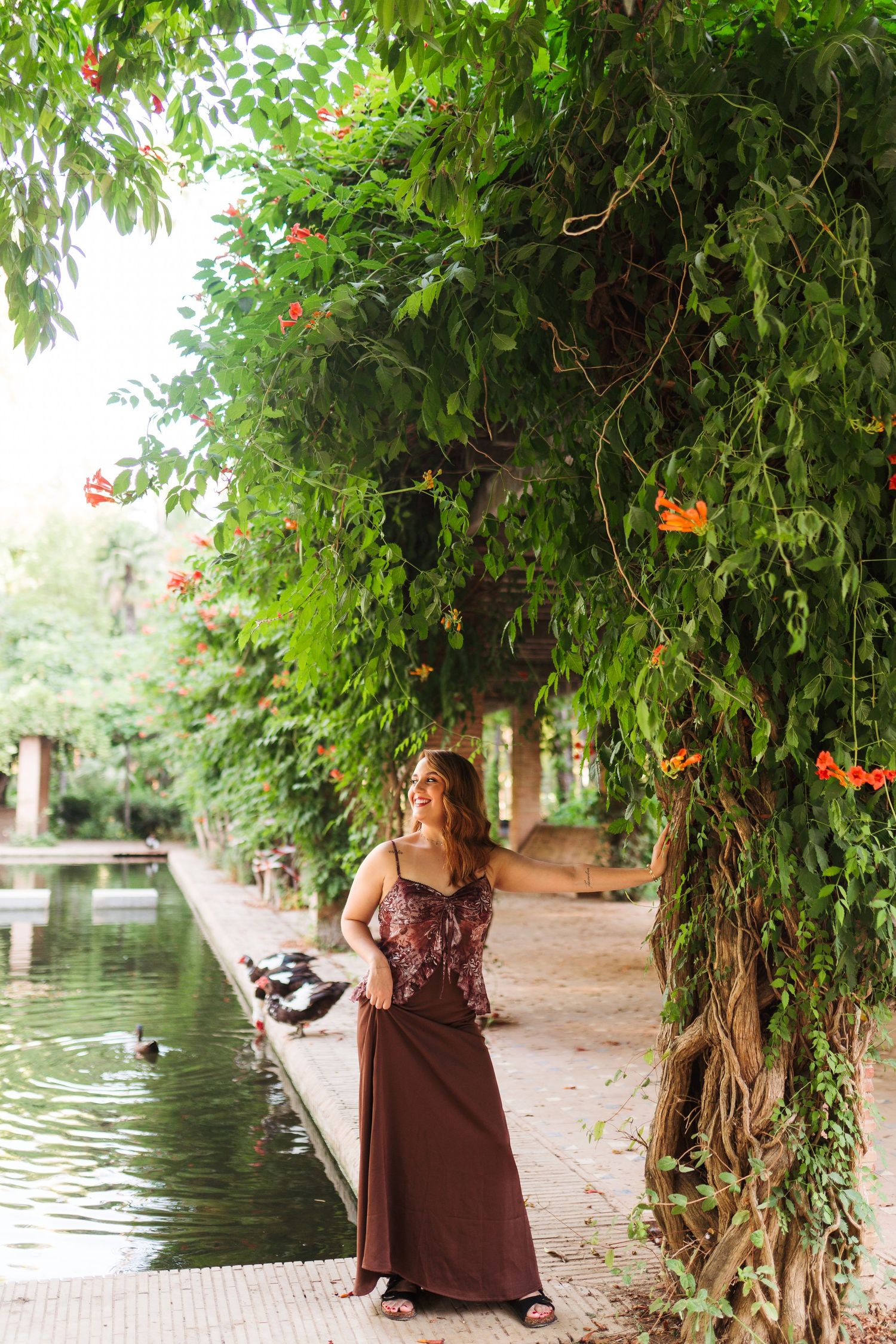 Woman in brown maxi dress standing by a tranquil pond surrounded by lush green trees with orange flowers.