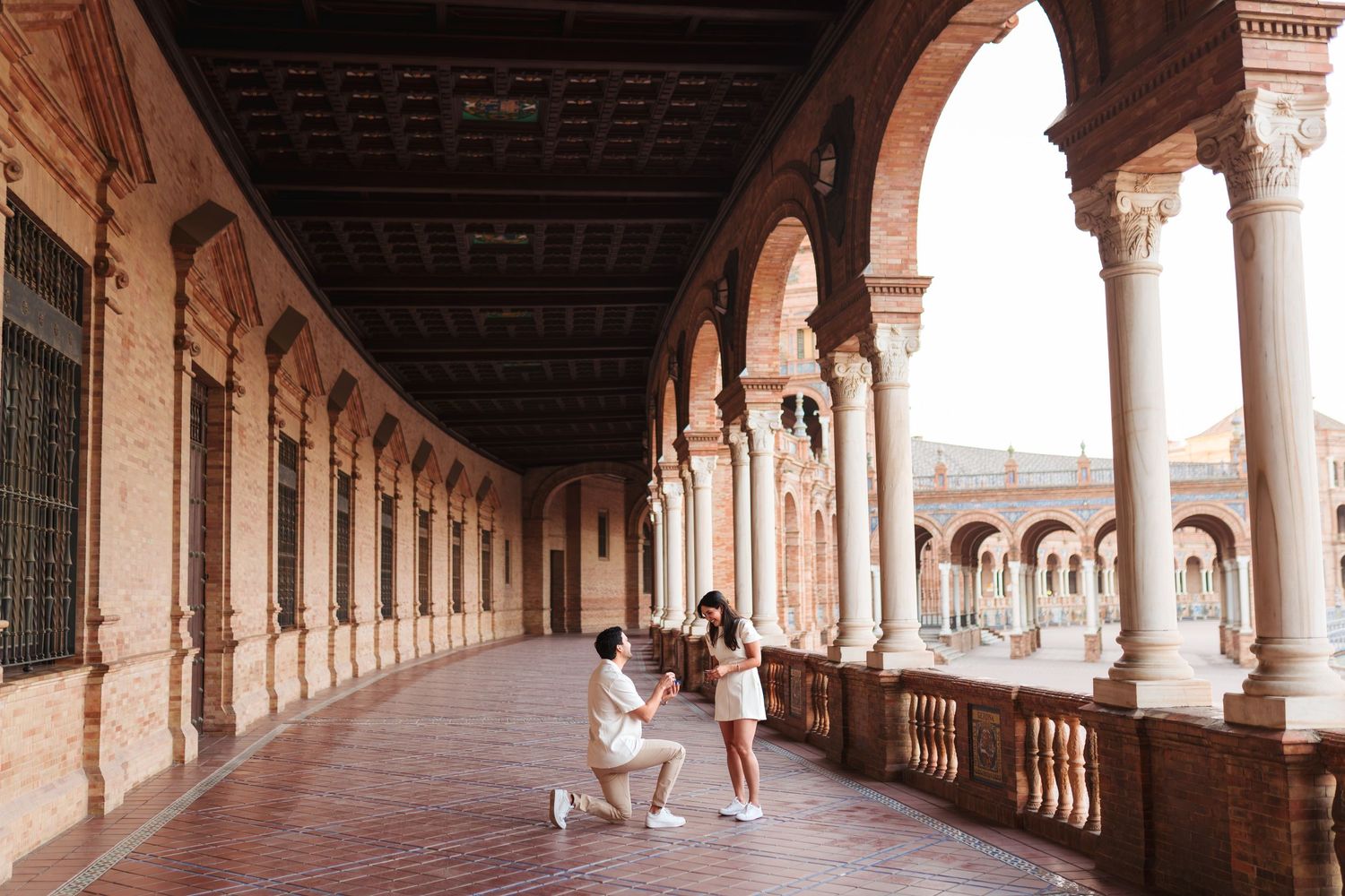 Man proposing to woman on one knee in ornate Spanish colonial arcade with arched columns and brick architecture.