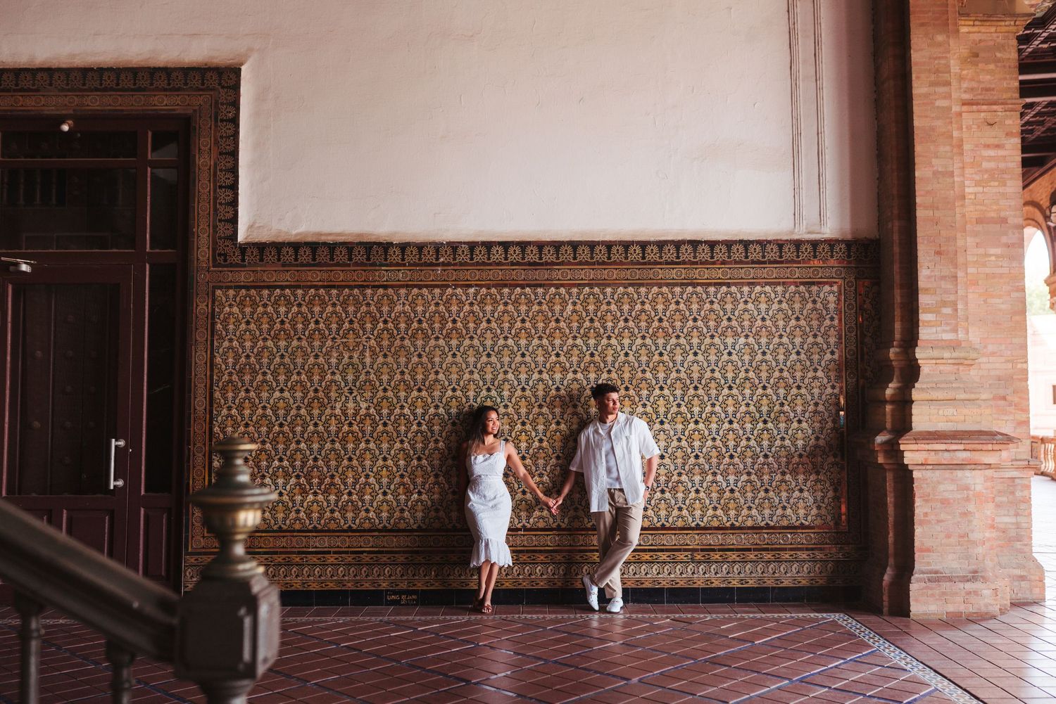 Couple holding hands walking in an ornate Spanish-style building with decorative tile walls and terracotta floors.