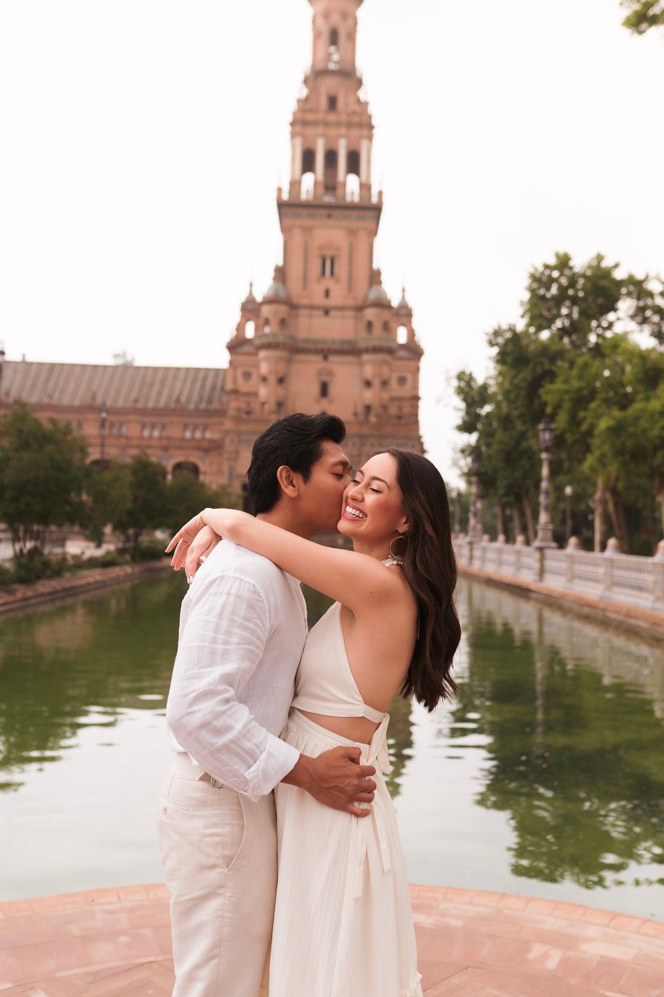 Happy couple sharing a romantic kiss near a scenic canal with a historic tower backdrop.