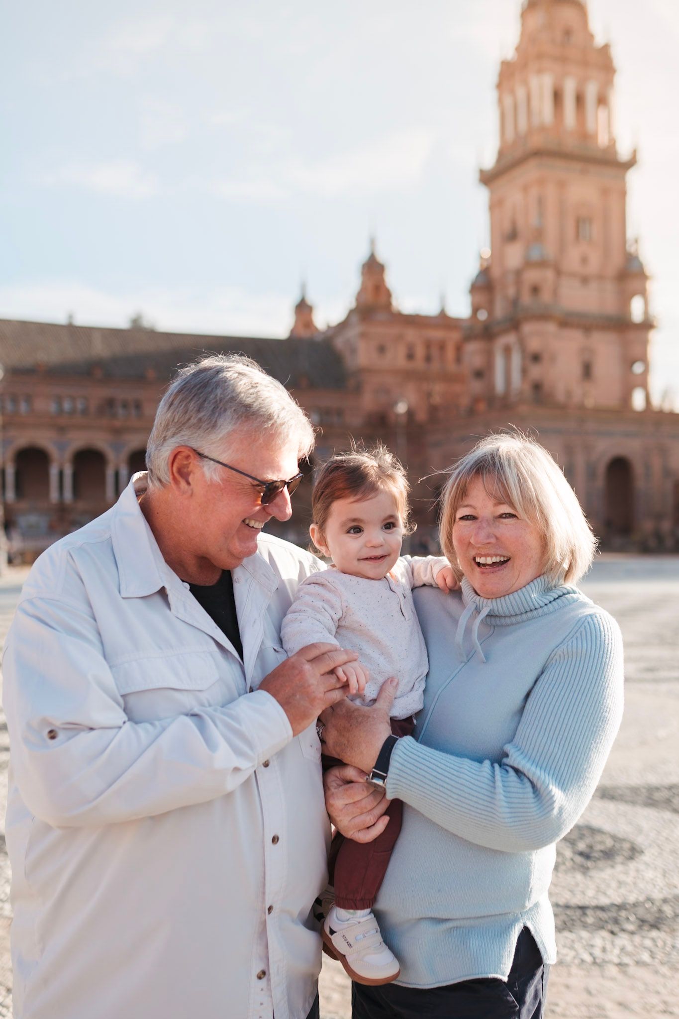 Smiling grandparents hold a baby girl in front of the iconic tower at Plaza de España, Seville.