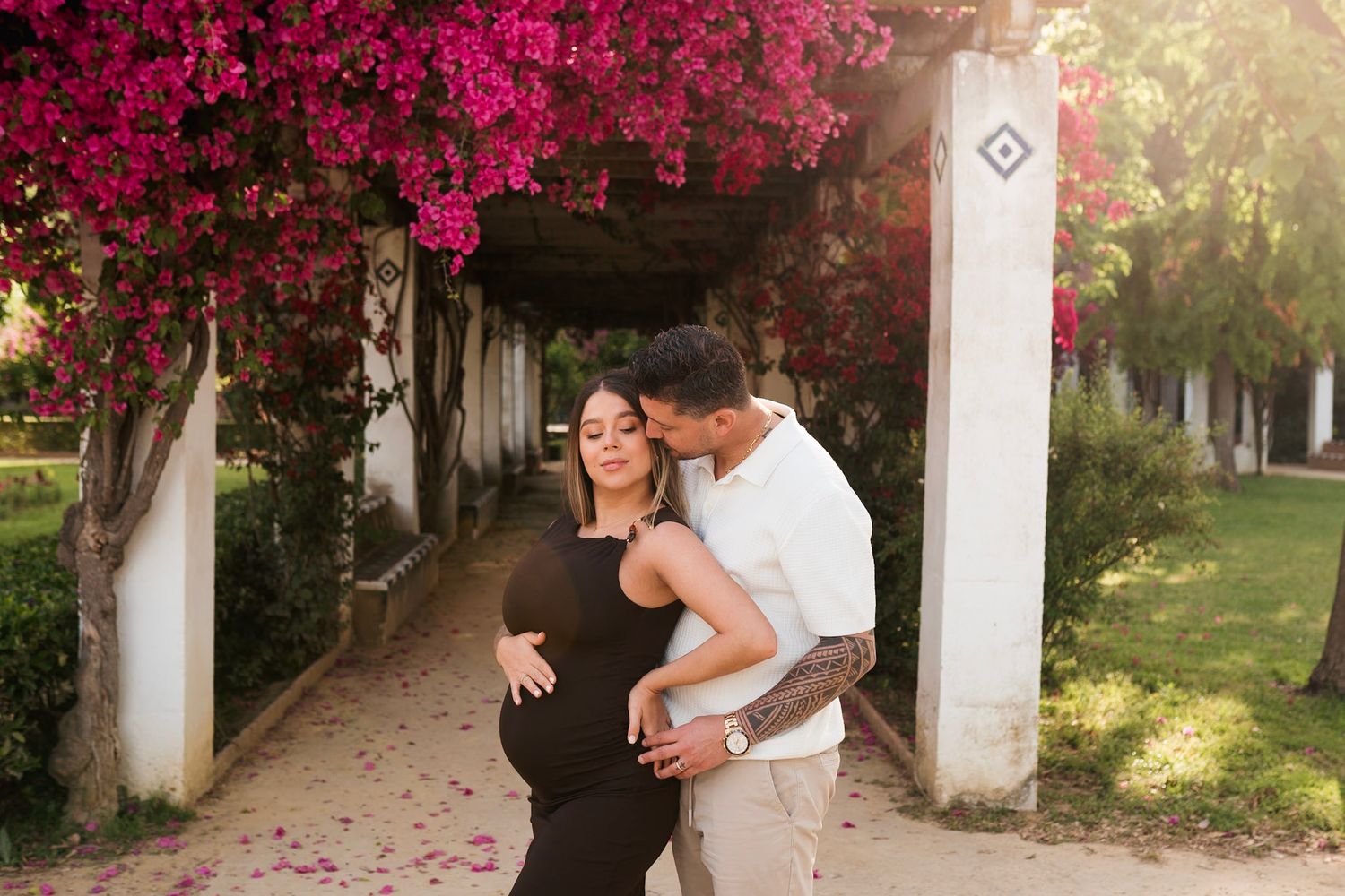 Pregnant woman in black dress embraced by tattooed partner under pink bougainvillea archway.