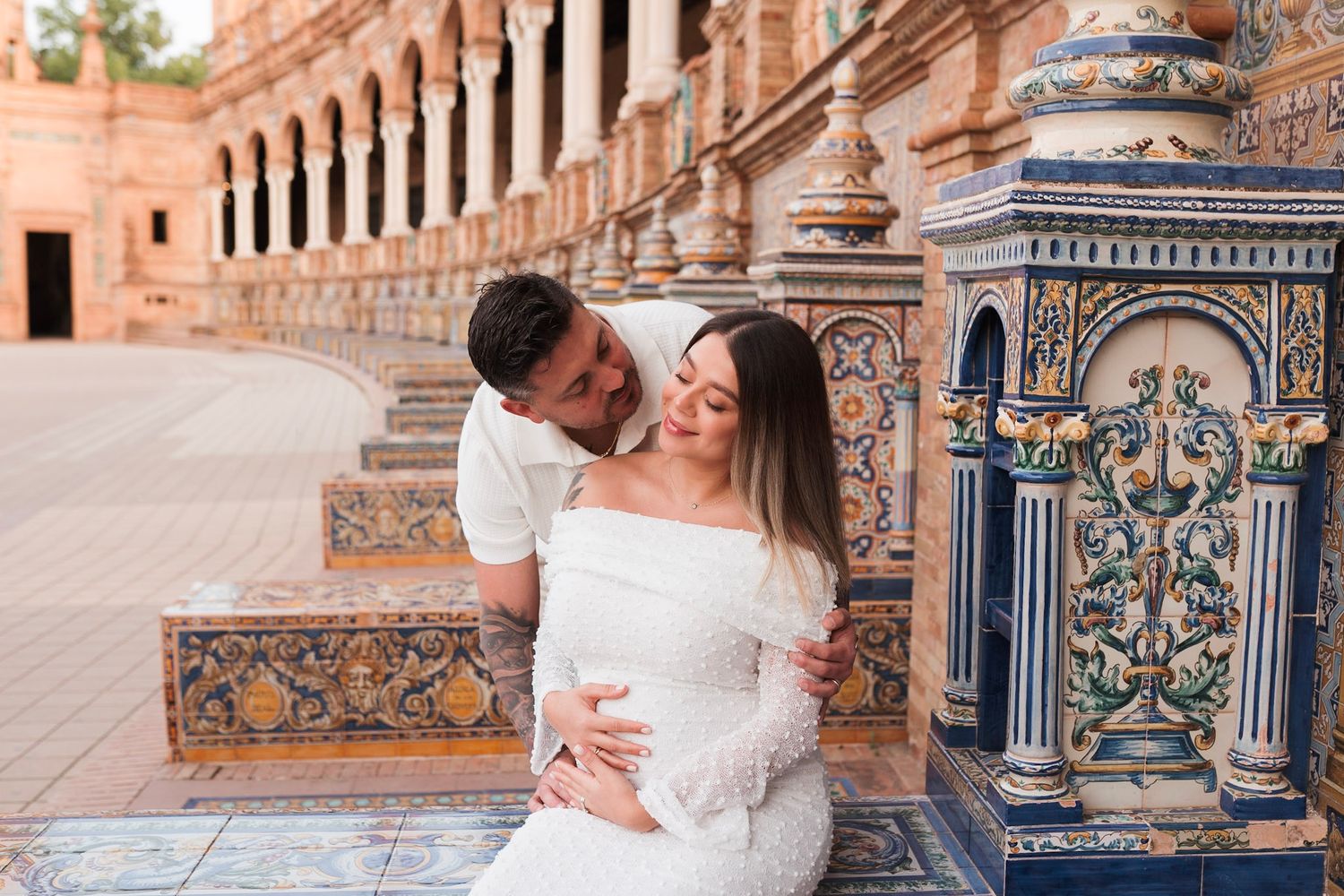 Couple embracing by ornate tiled column at Plaza de España Seville, woman in white maternity dress.