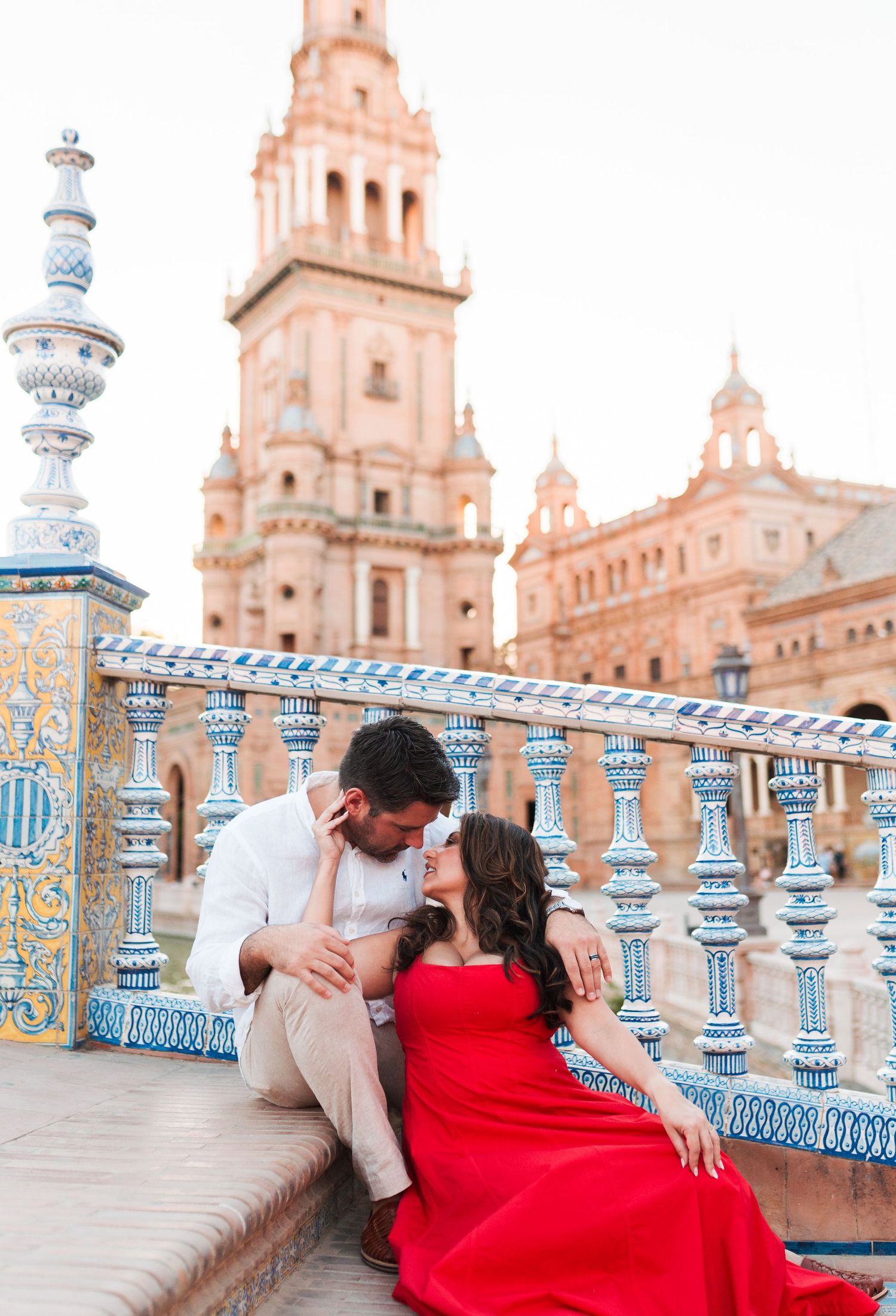 Romantic couple embracing at Plaza de España in Seville, woman in red dress, ornate tiled balustrade and tower backdrop.