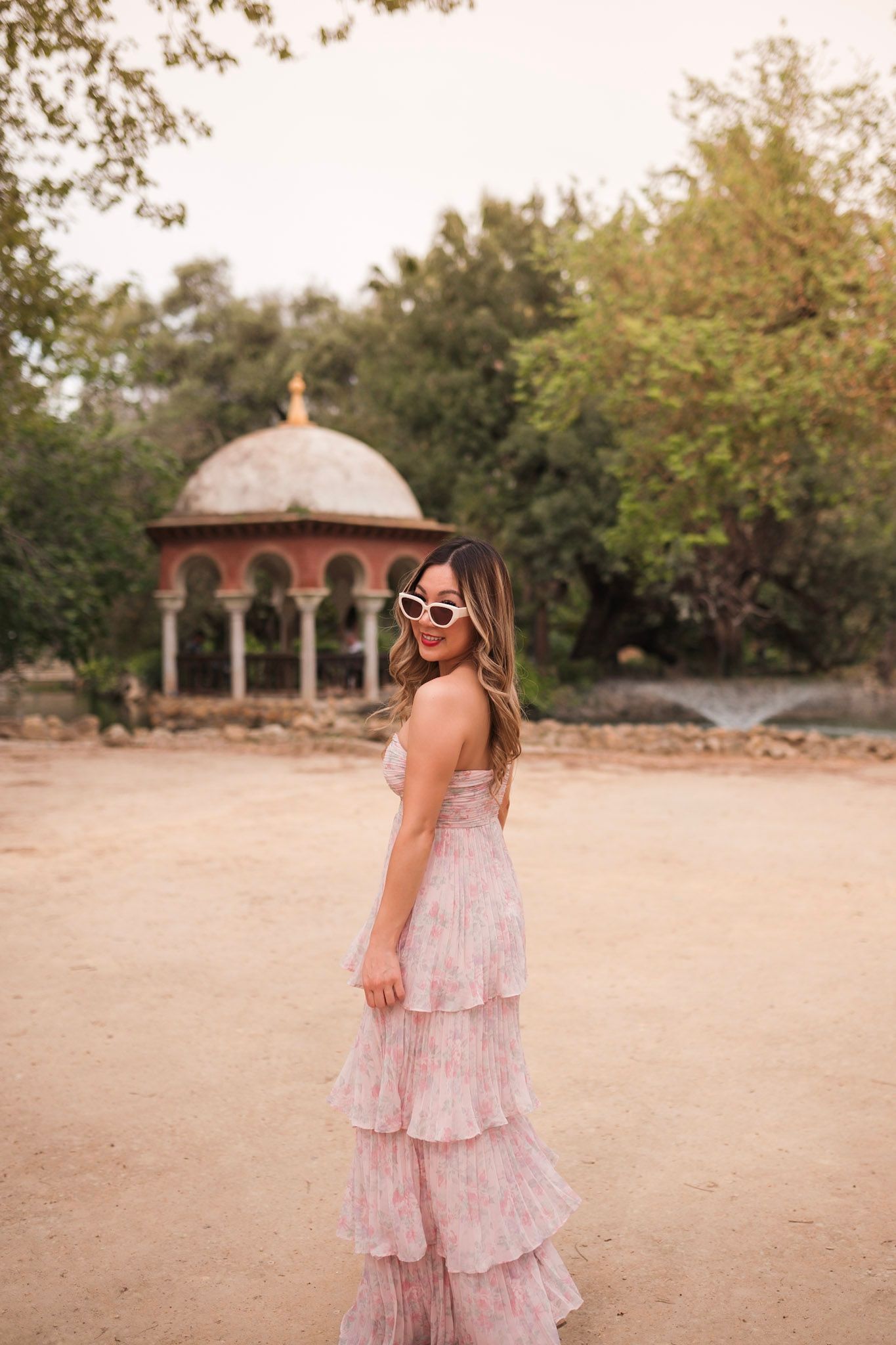 Woman in pink ruffled maxi dress with sunglasses poses near historic domed pavilion.
