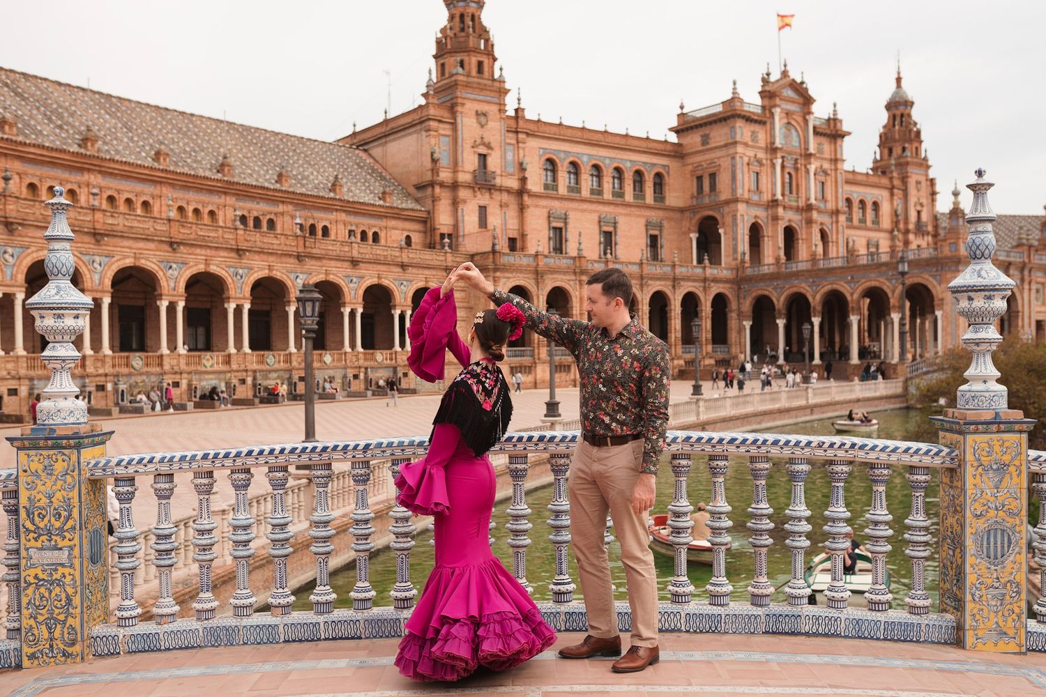 Couple dancing flamenco at Plaza de España Seville, woman in pink ruffled dress twirling near ornate tiled bridge.