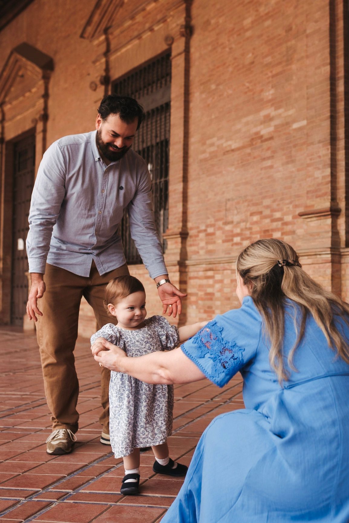 A family with a toddler girl in a floral dress taking first steps between parents at a brick building.