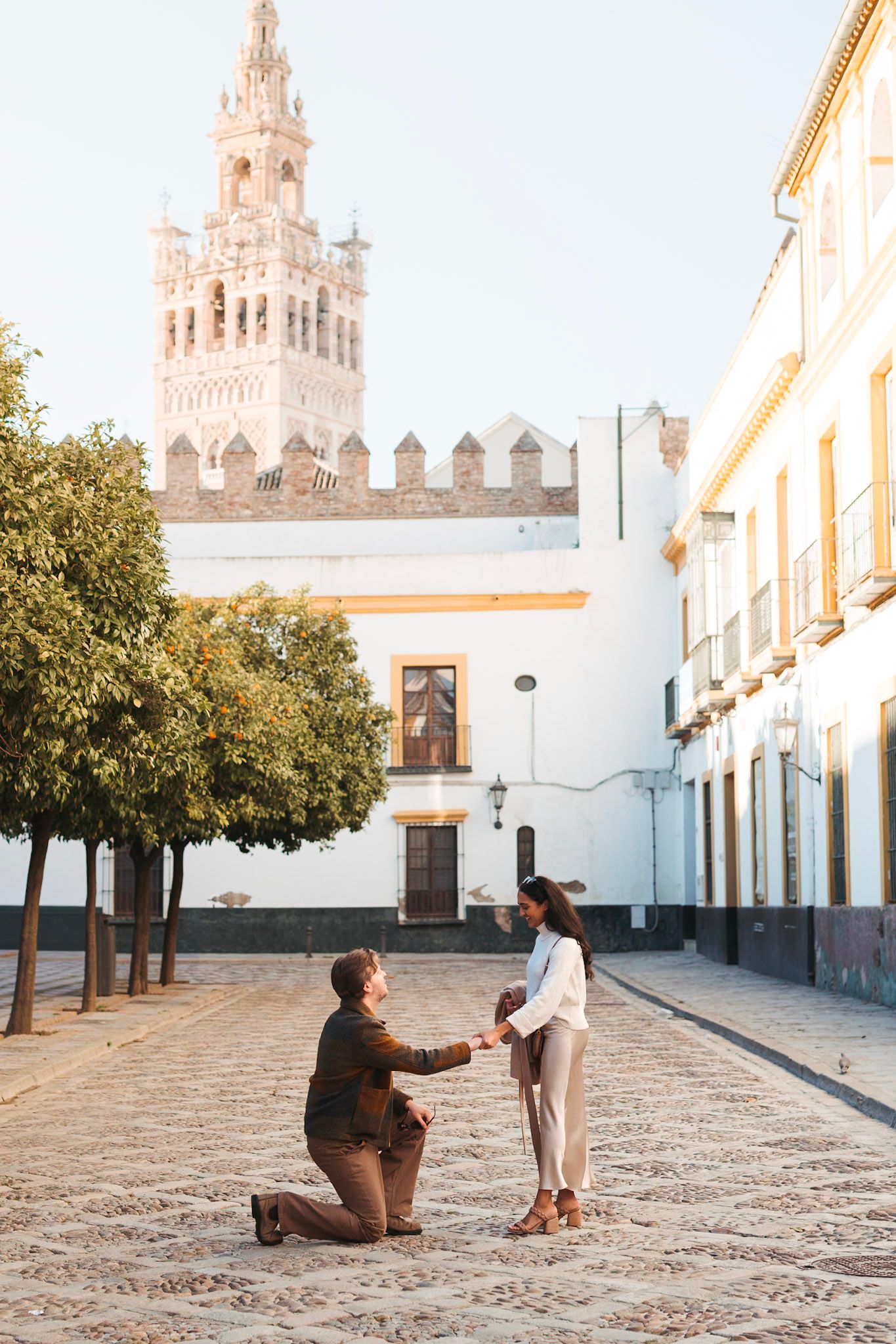 Man on one knee proposing to woman in Seville street with Giralda tower visible in background.