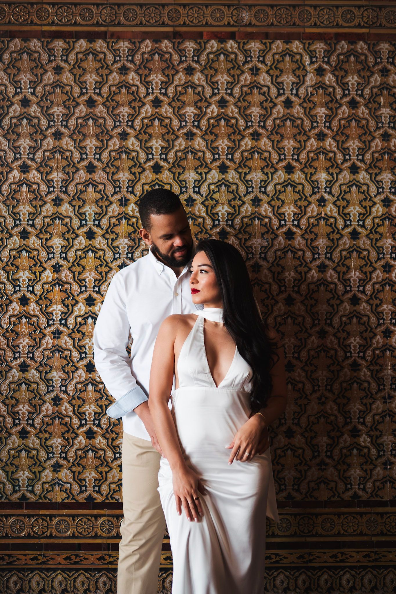 Elegant couple posing before ornate decorative tilework wall, woman in white dress with red lips.