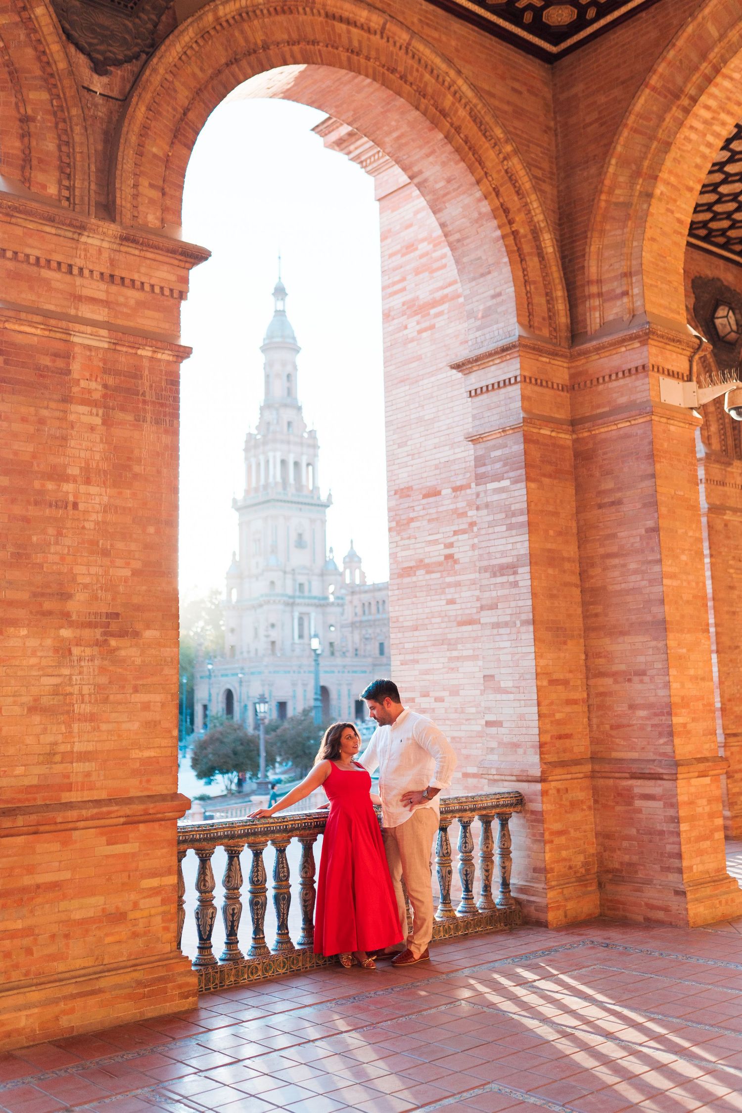 Couple in red dress and white shirt embracing under brick arches at Plaza de España, Seville, Spain.