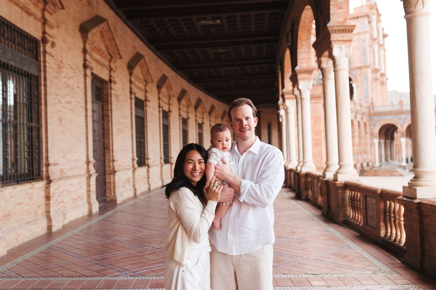 A couple holds their baby while posing in an elegant Spanish arcade with ornate columns and warm terracotta tones.