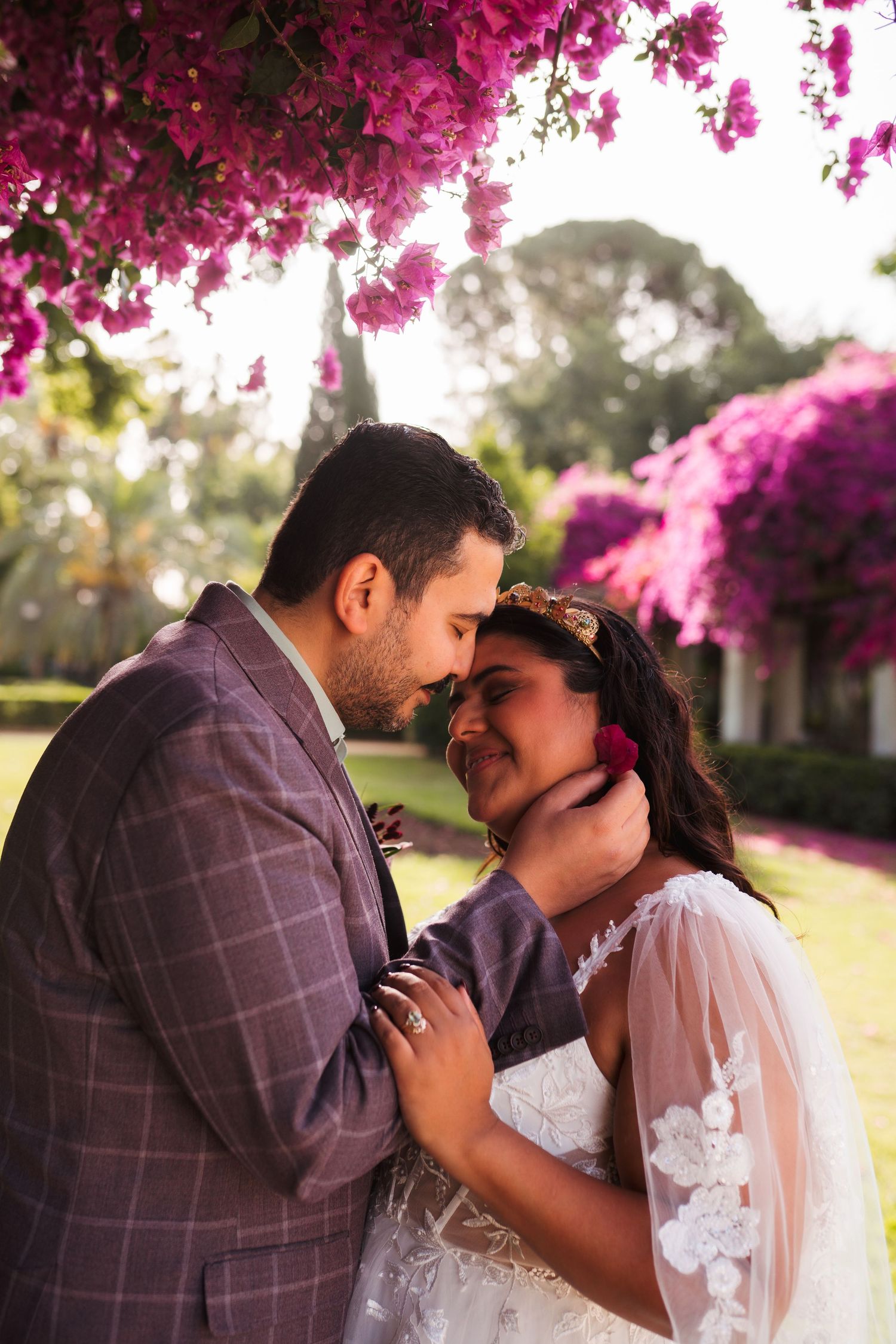 Bride and groom share a romantic moment under vibrant pink bougainvillea blooms in a lush garden setting.
