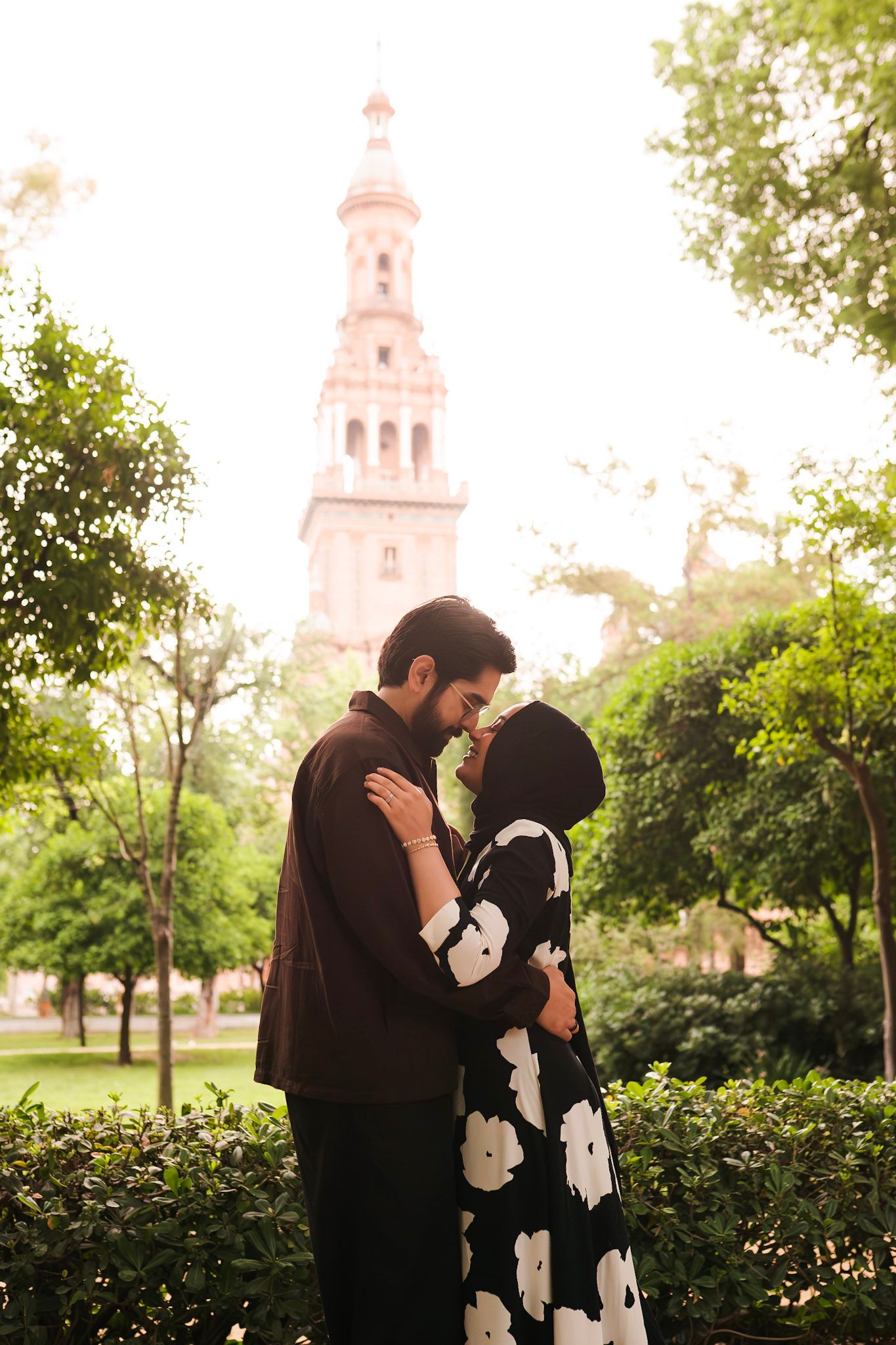 Couple embracing in lush garden with Plaza de España Seville tower visible through trees behind them.