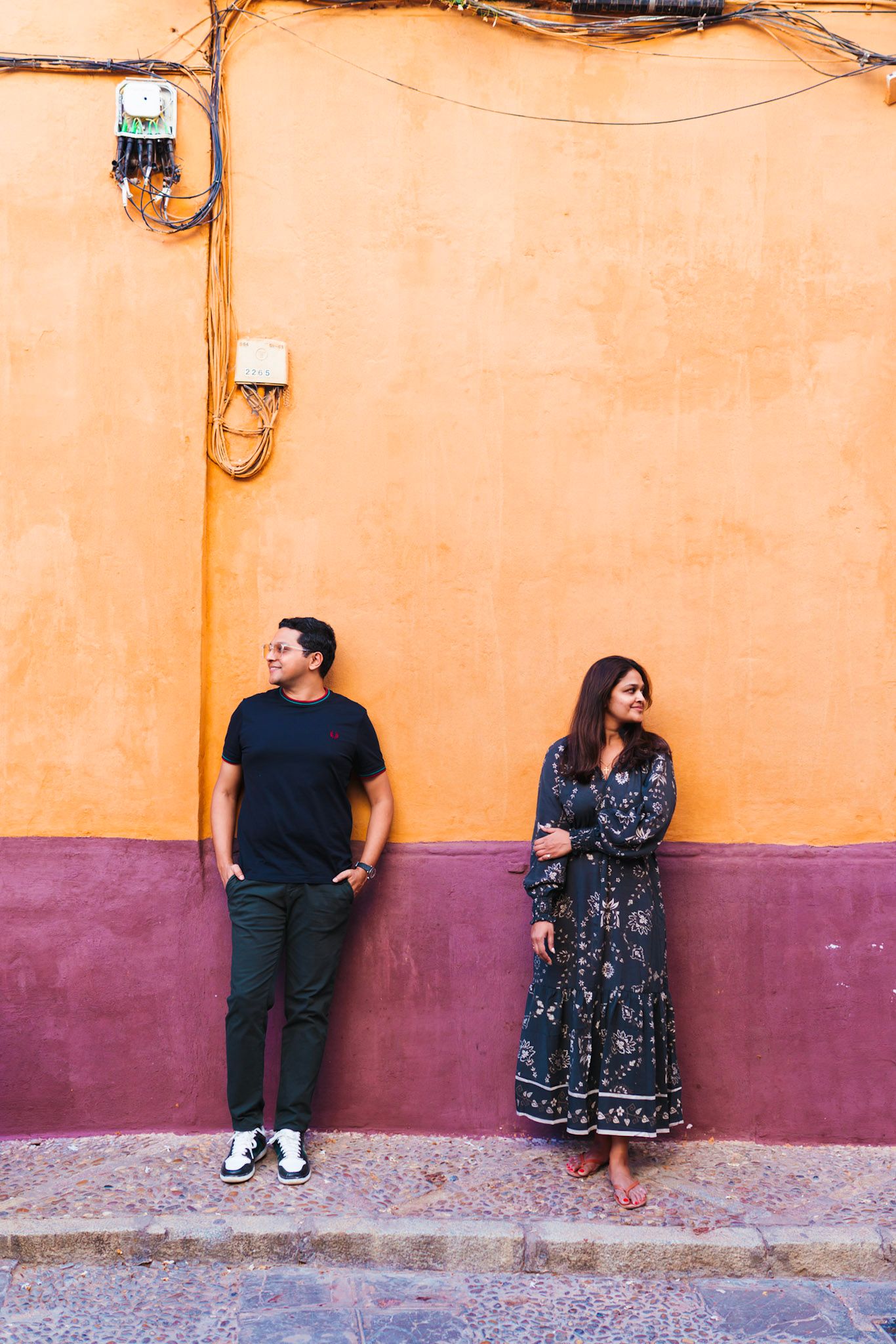 Couple standing apart against a colorful orange and purple wall on a cobblestone street in a vibrant urban setting.