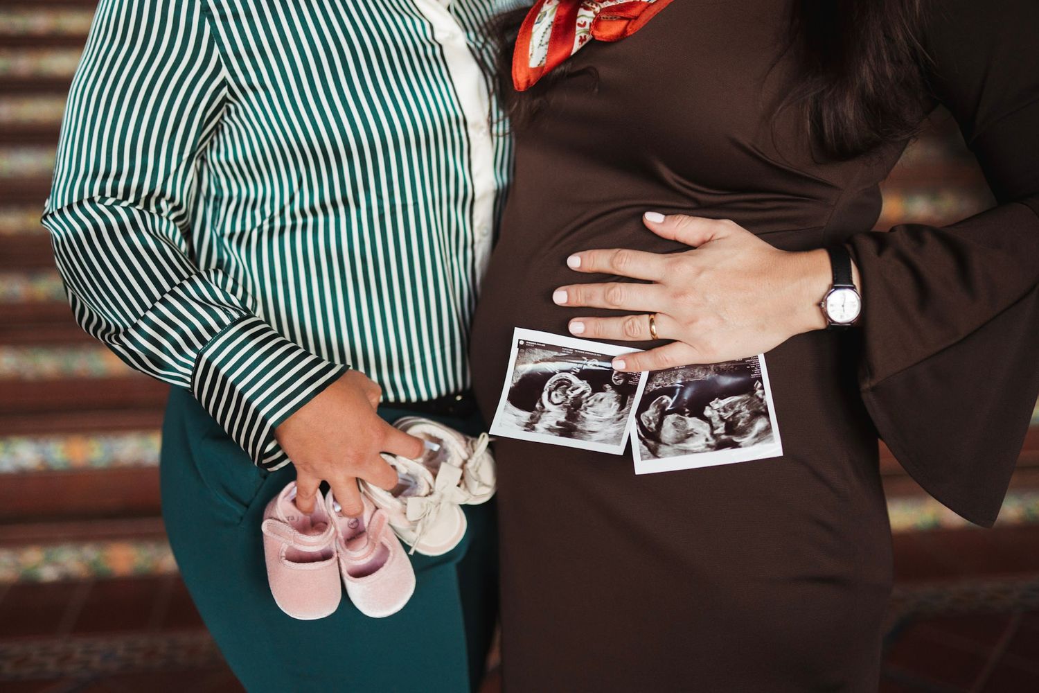 Same-sex couple holding ultrasound photos and pink baby shoes over pregnant belly.