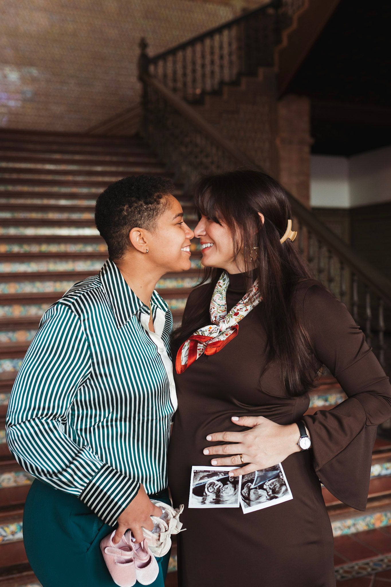 Same-sex couple nearly kissing on ornate tiled staircase during maternity photo session.