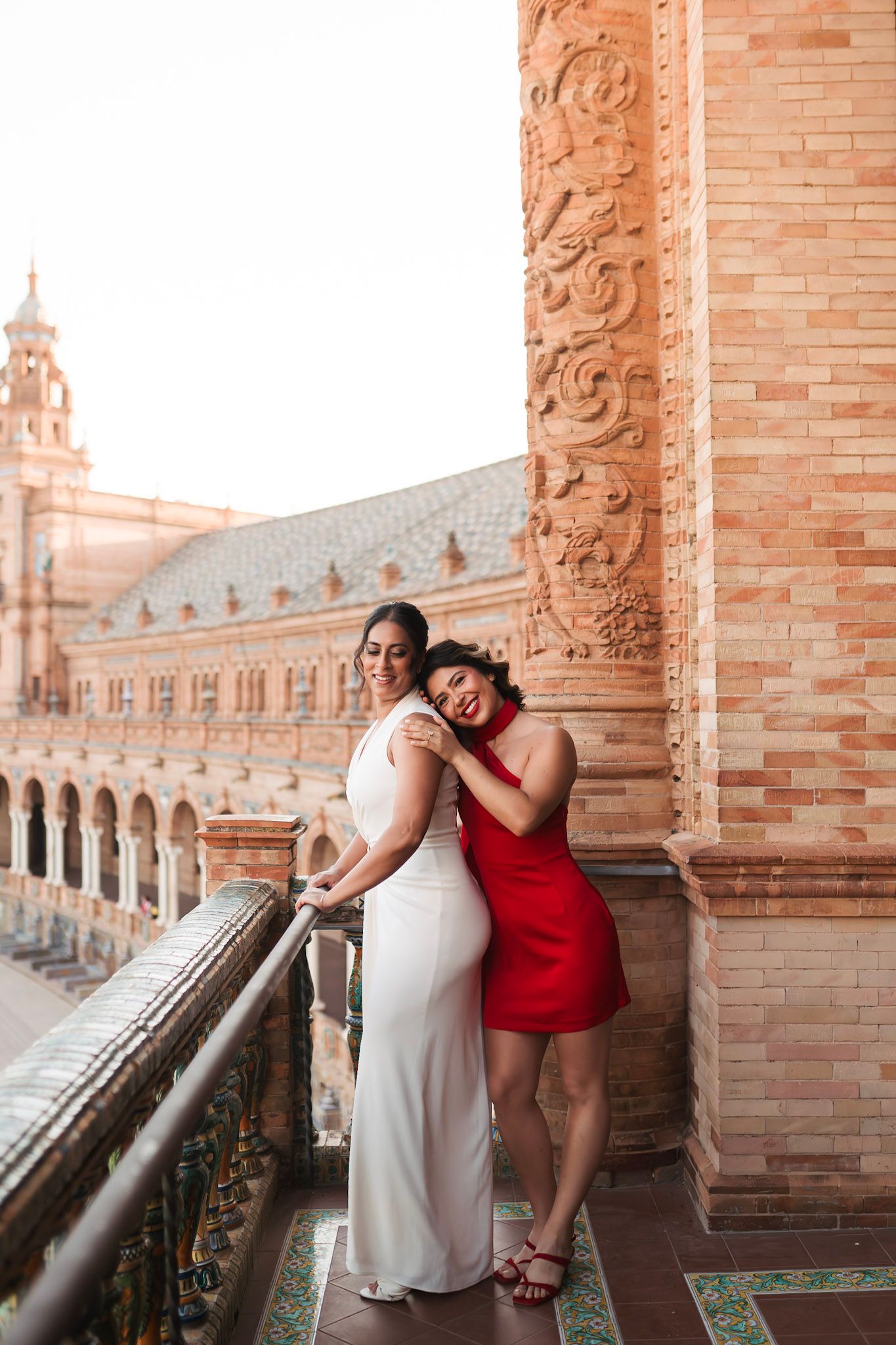 Two women embracing on balcony at Plaza de España, one in white, one in red.