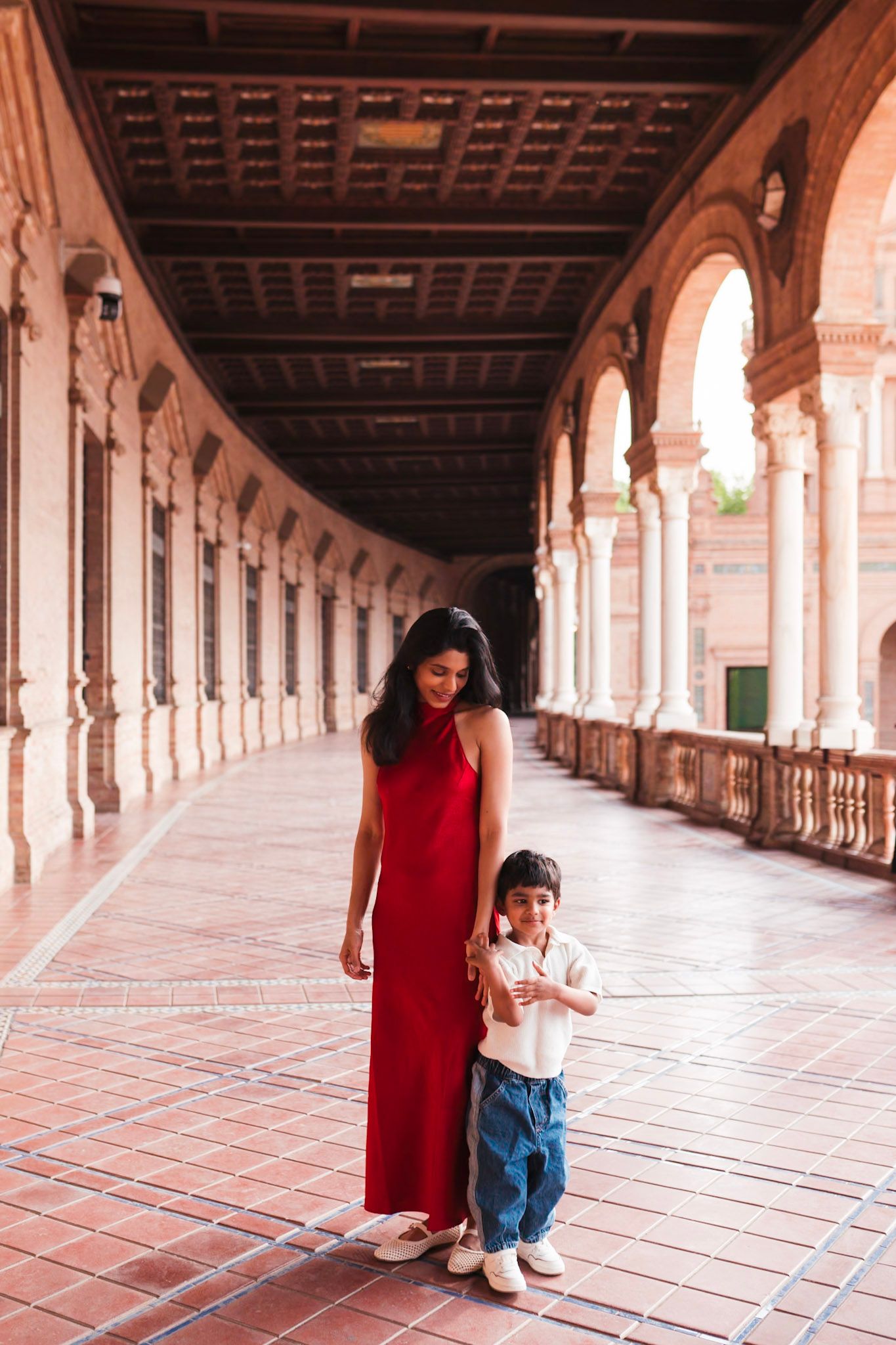 A mother in a red maxi dress walks with her young son through an arched corridor in Seville Spain.