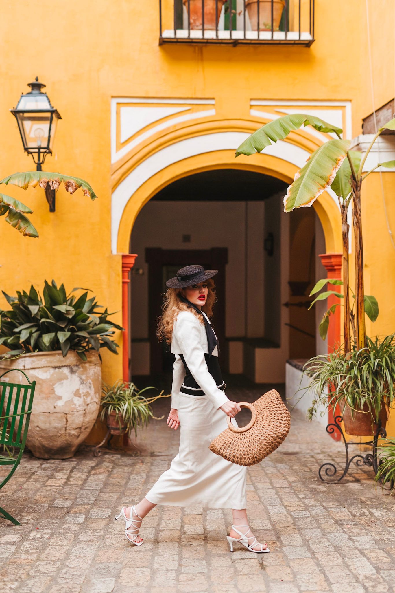 Elegant woman in white skirt and black hat walks through yellow courtyard with tropical plants in Seville.