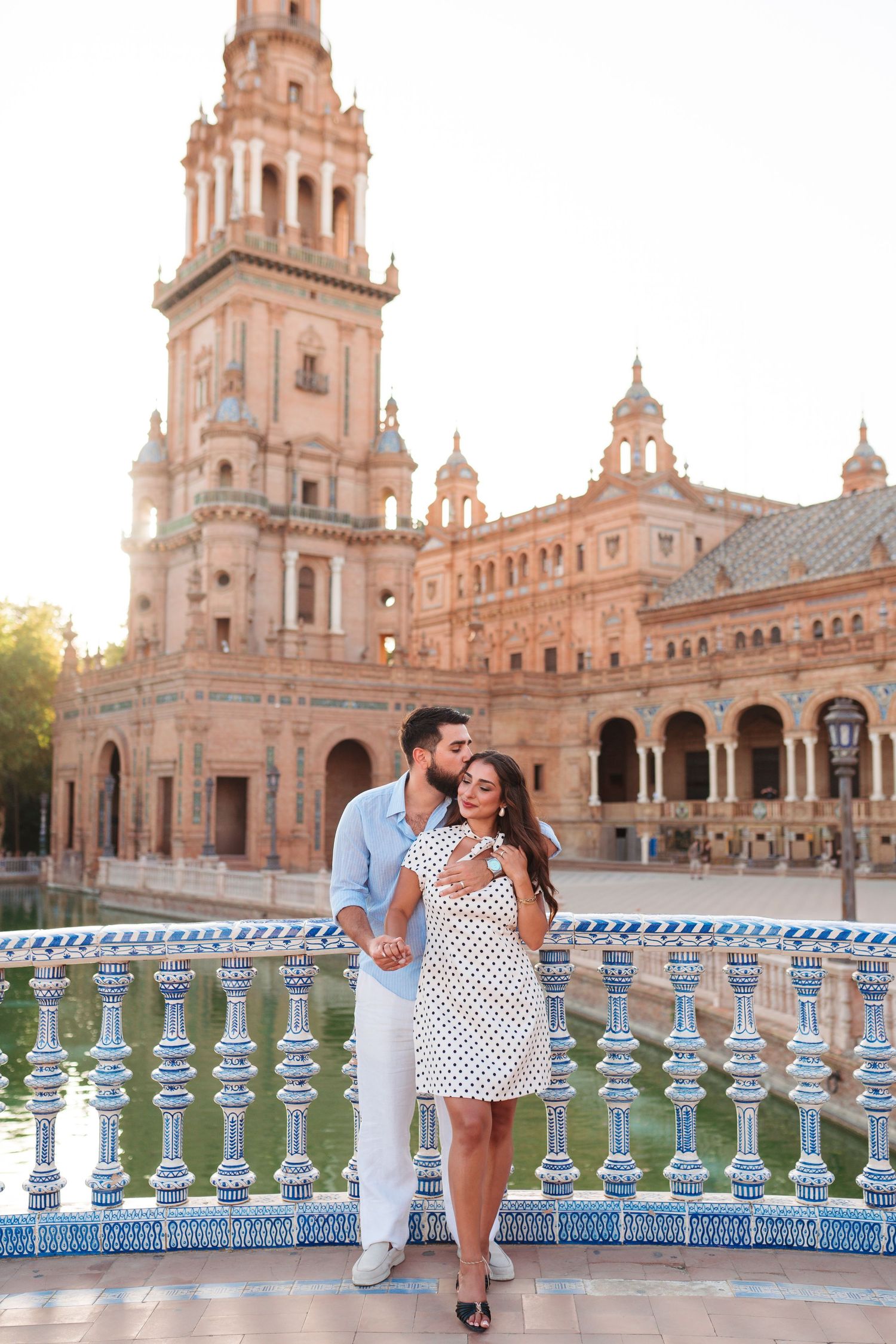 Romantic couple embracing at Plaza de España in Seville, Spain, with ornate baroque architecture and blue tiled bridge.