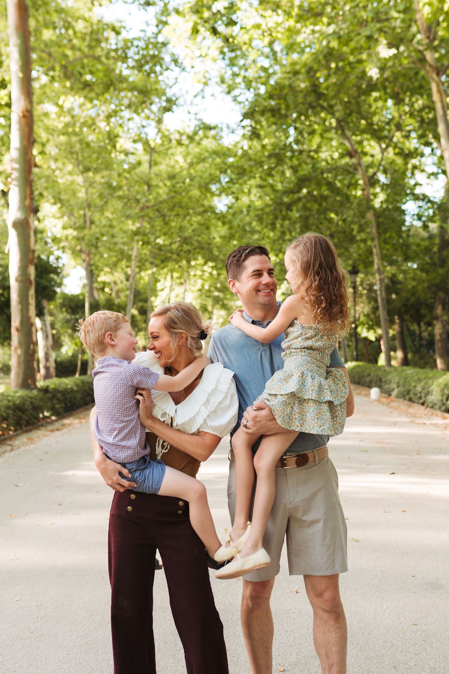 Happy family of four enjoying a sunny day in a tree-lined park, parents holding young children while smiling.