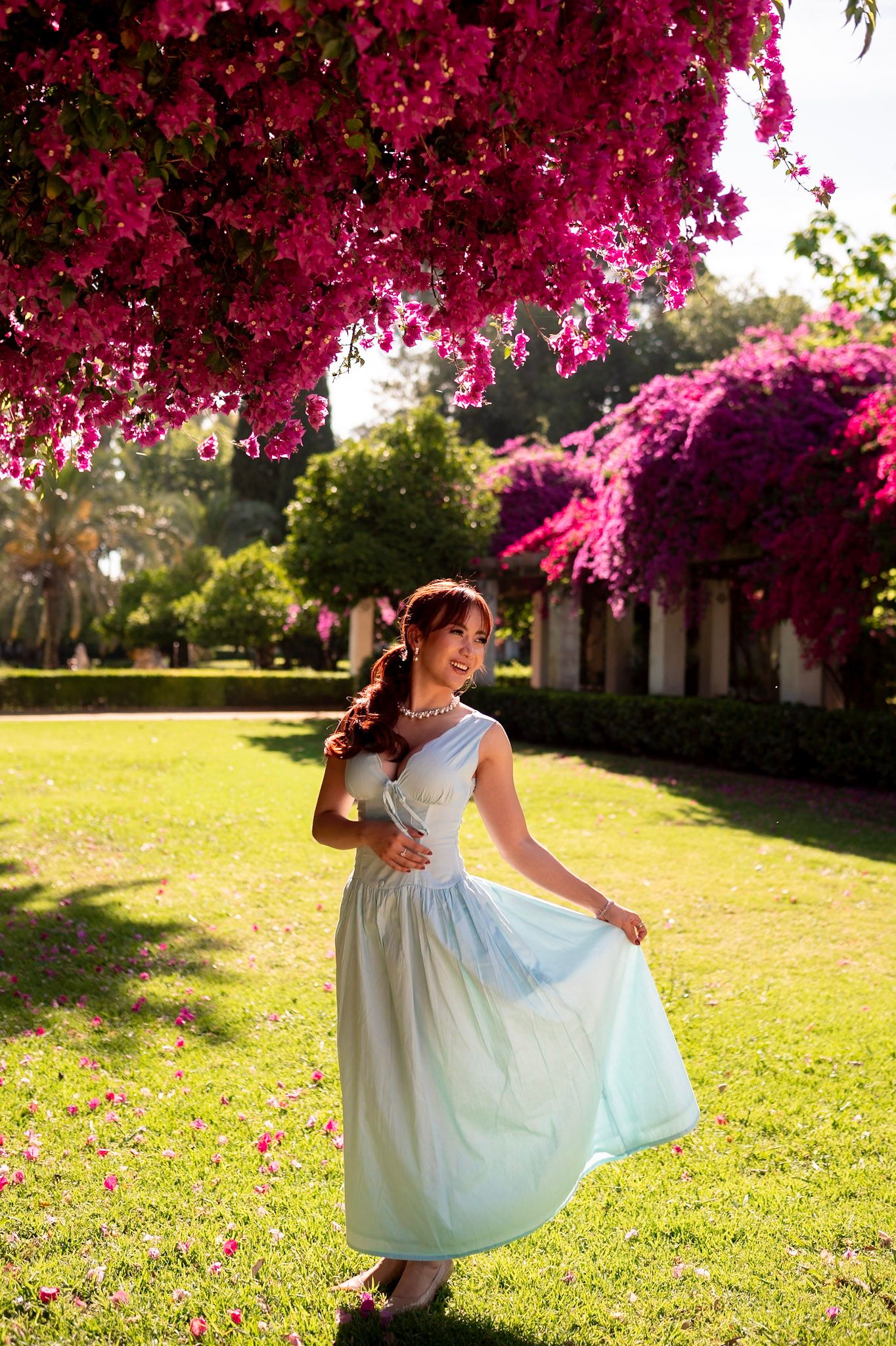 Smiling woman in white maxi dress twirls under vibrant pink bougainvillea trees in a sunny garden park.