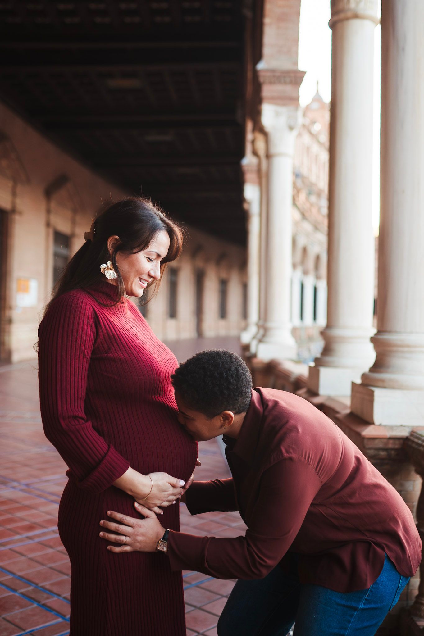 Pregnant woman in red dress smiles as partner kisses her baby bump in elegant columned arcade.