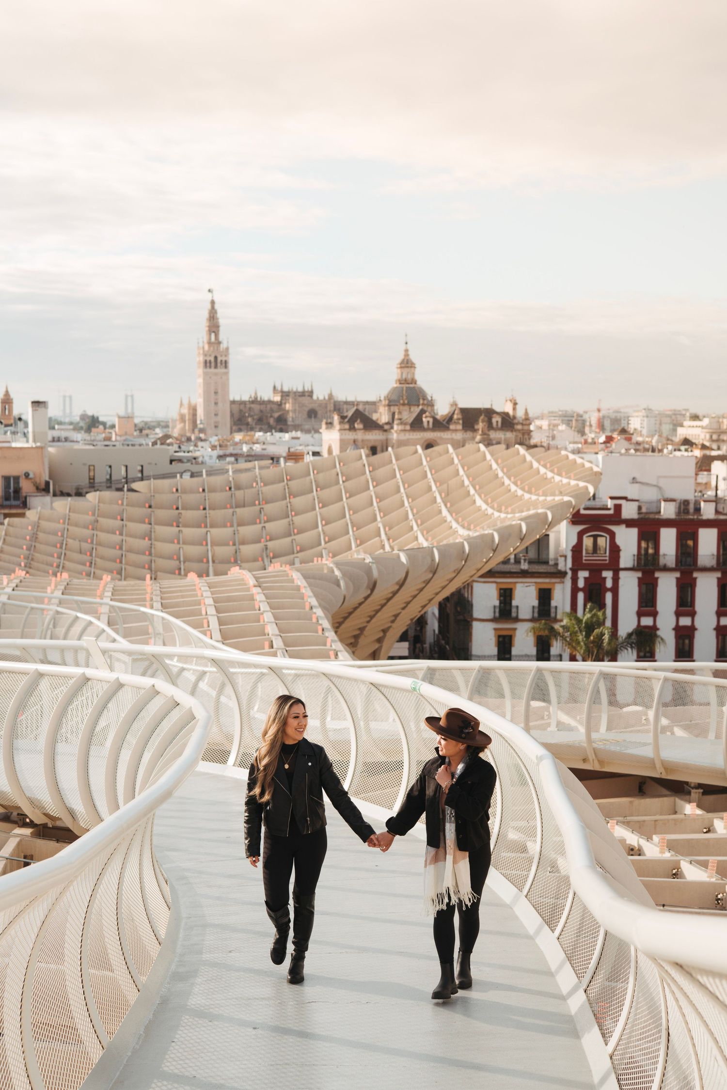 Two women holding hands walking on the curved Metropol Parasol walkway with Seville skyline at sunset.