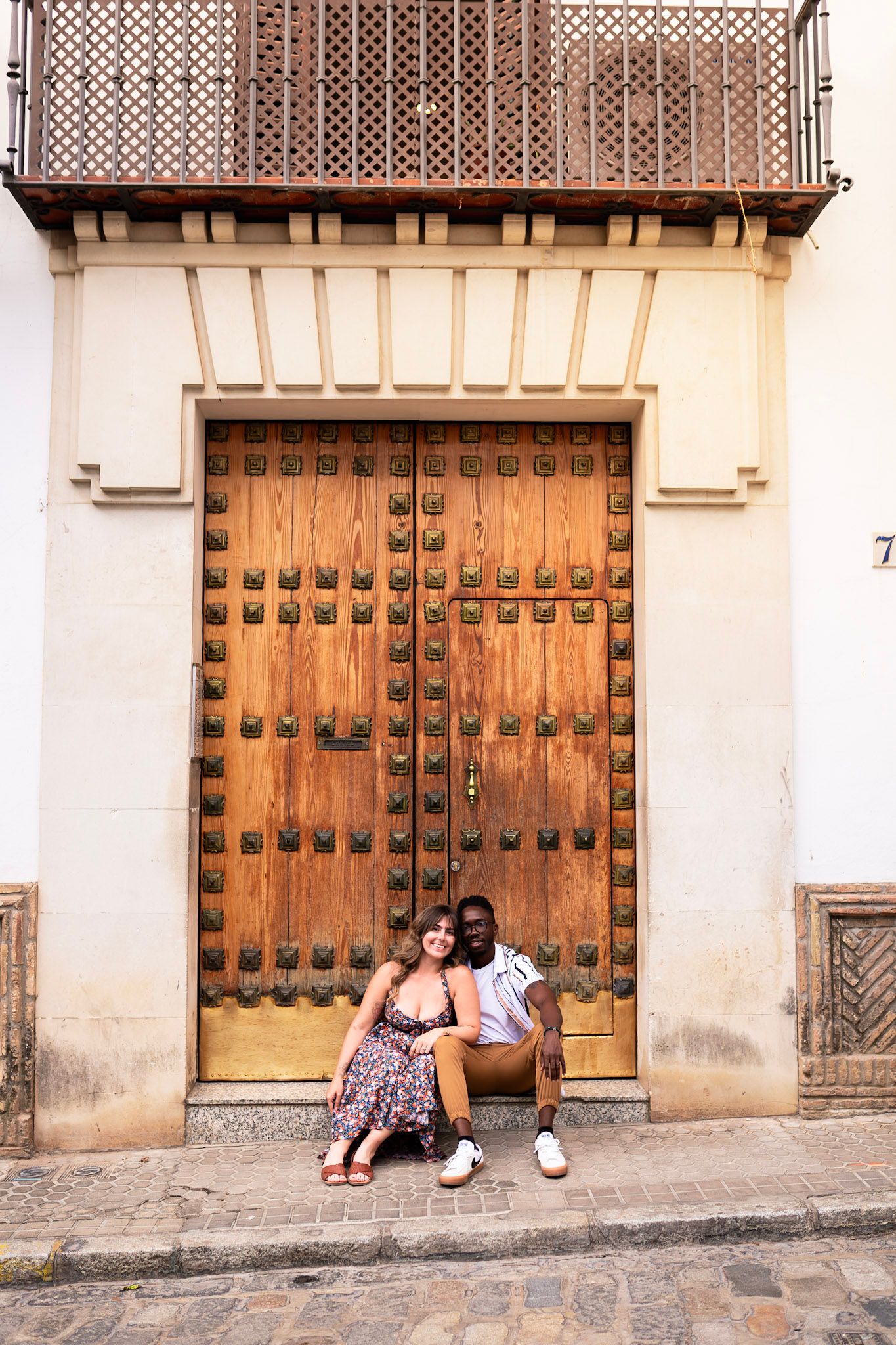 Smiling couple sitting together in front of a large ornate wooden door on a cobblestone street in Seville.