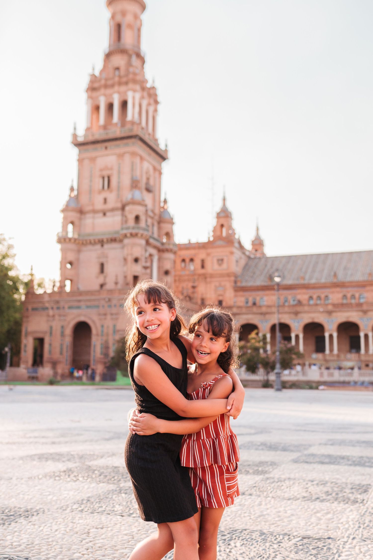 Two young girls hugging and smiling in front of Plaza de España tower in Seville, Spain.