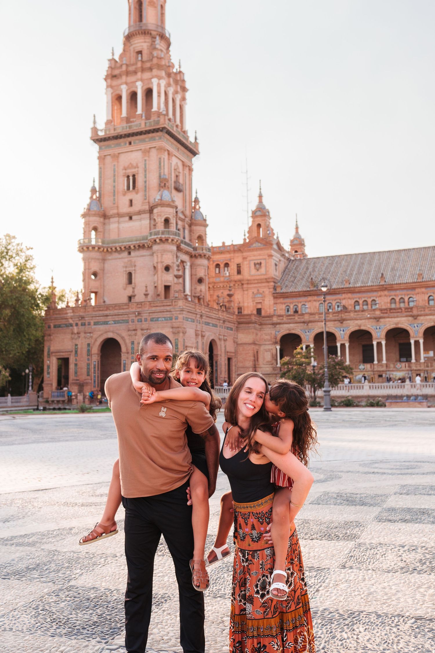Family of four posing happily in front of Plaza de España tower in Seville, Spain, on a sunny day.