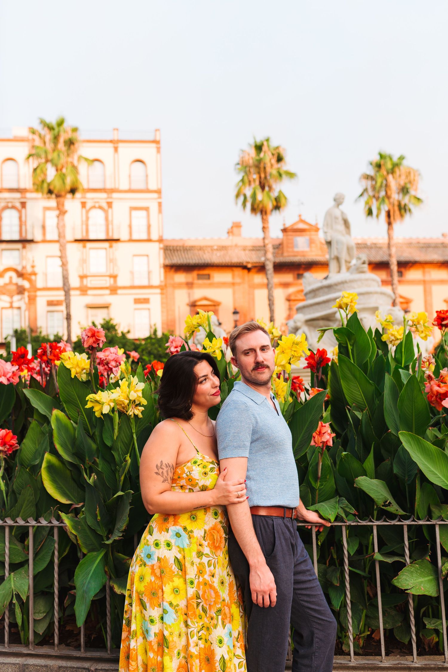 Couple embracing in front of colorful tropical flowers, palm trees, and a historic building at golden hour.