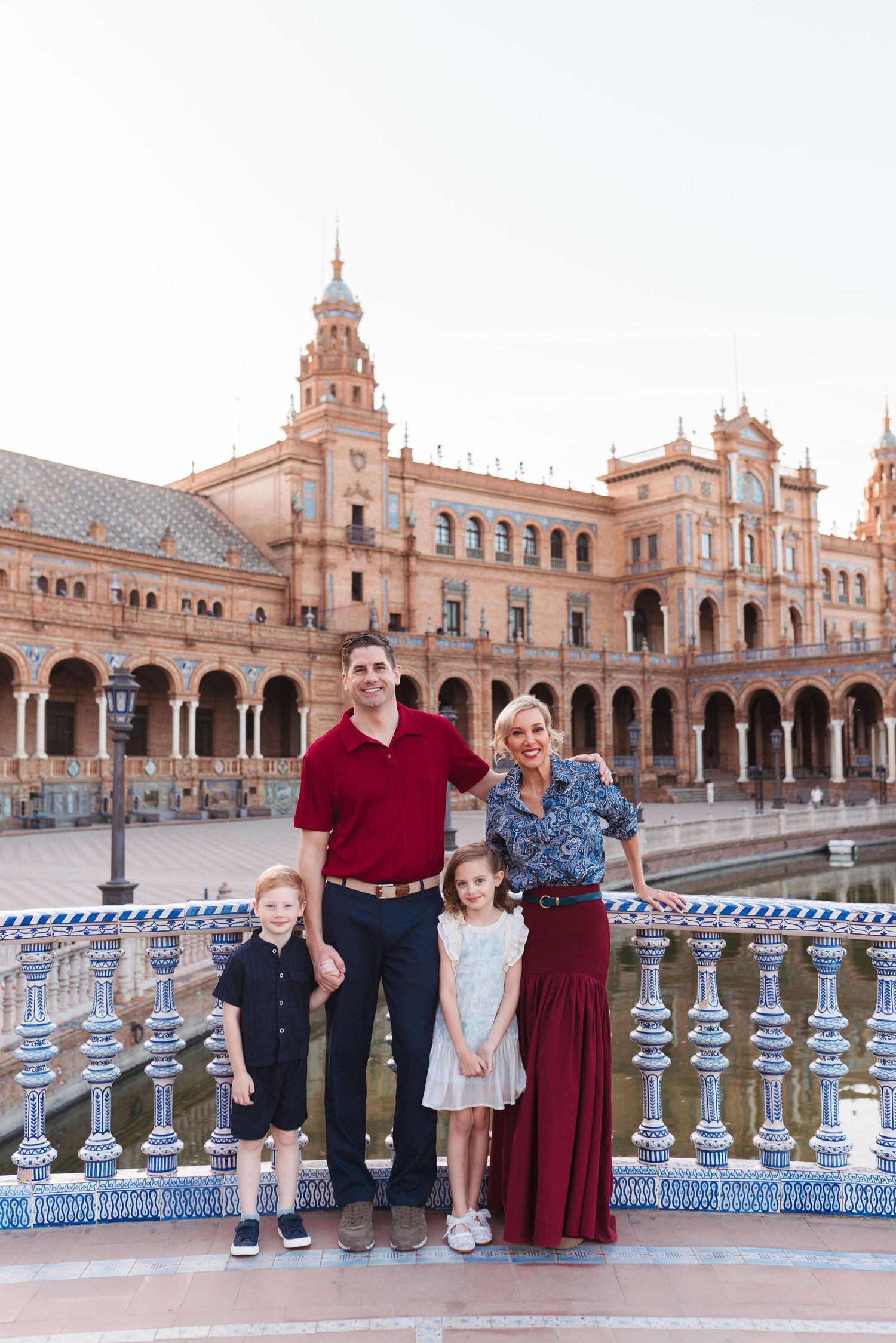 Family of four posing at Plaza de España in Seville, Spain, with ornate architecture and blue tiled bridge.