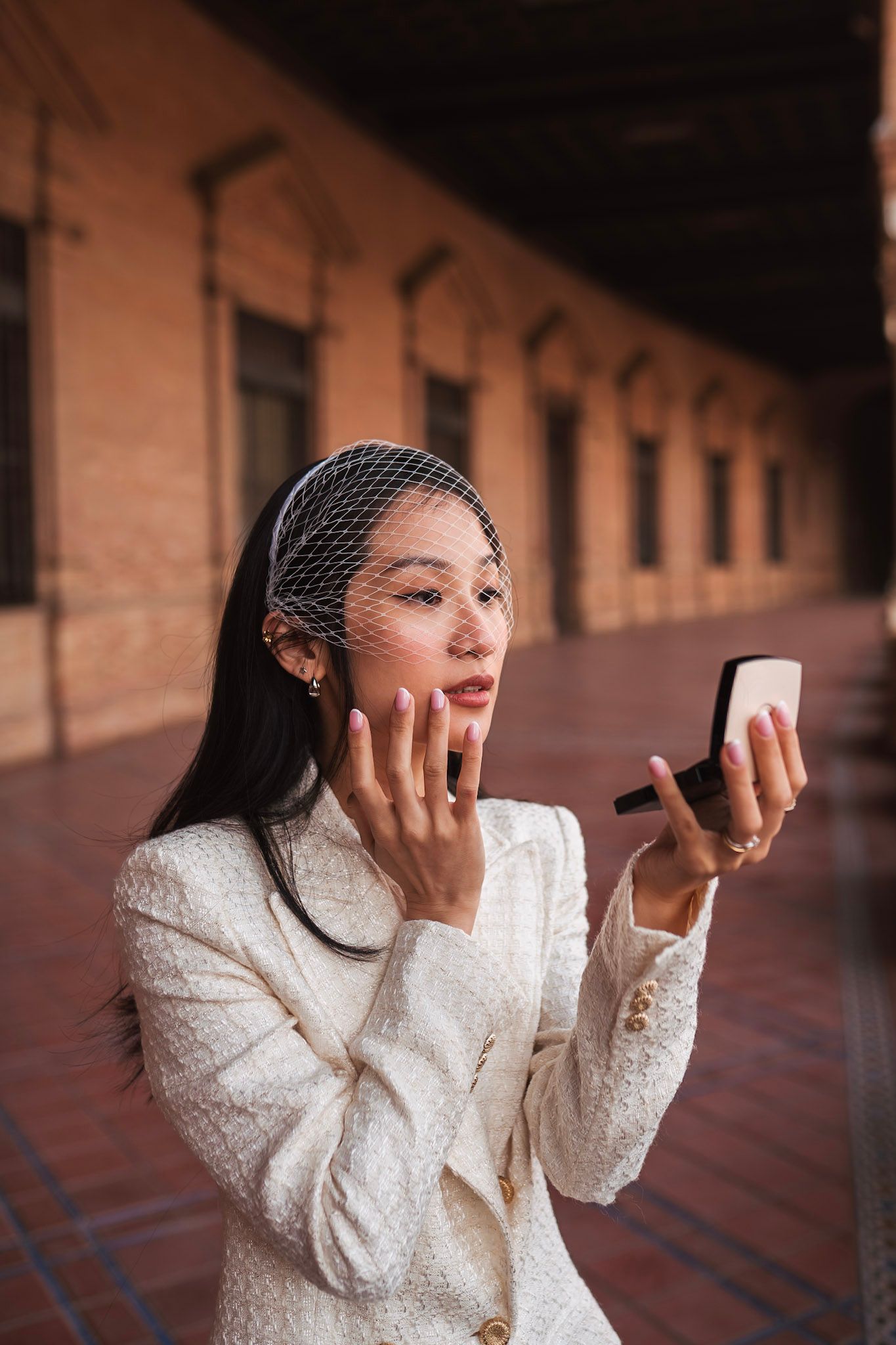 Woman in white jacket checking makeup in compact mirror at historic arcade.