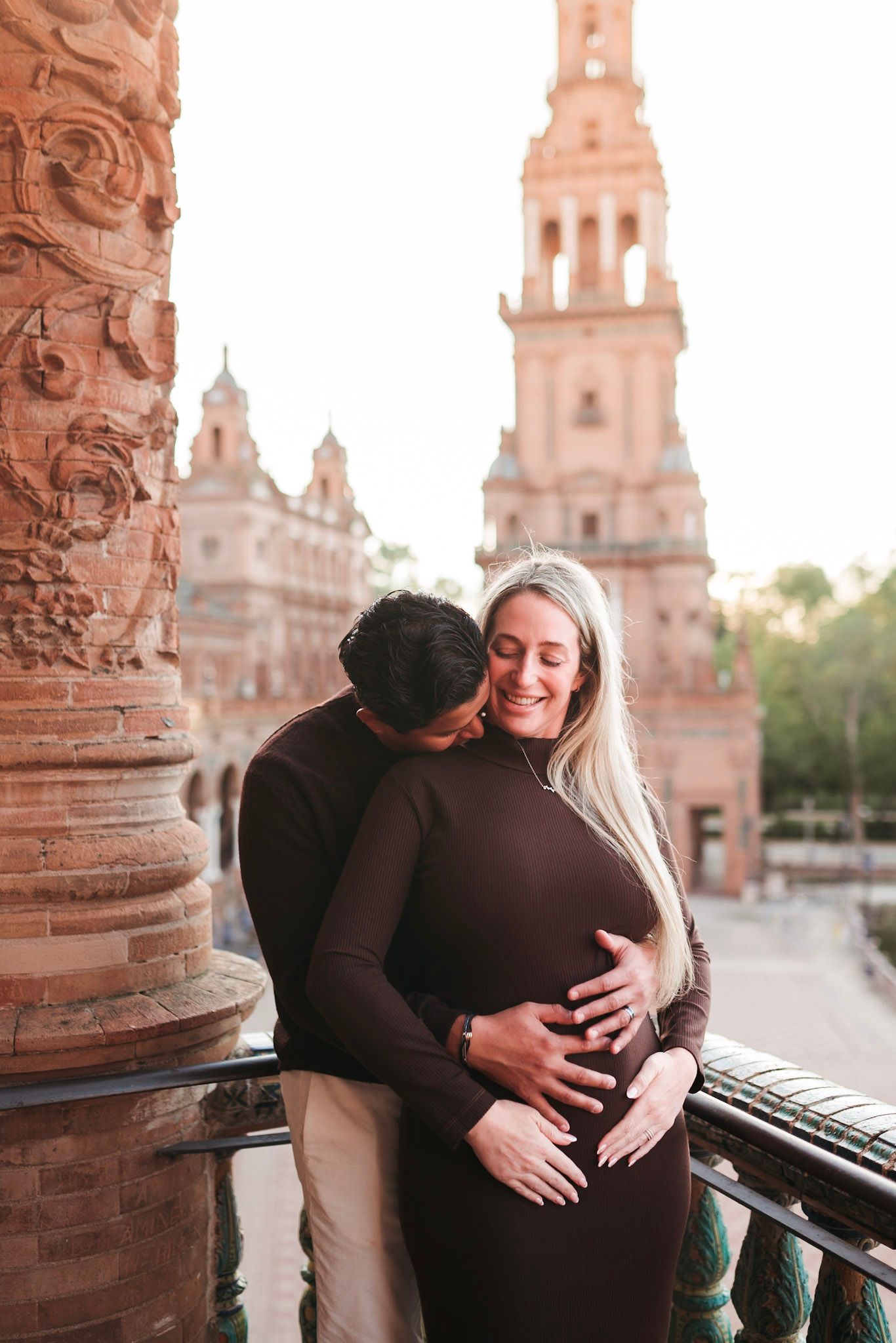 Couple sharing tender moment on balcony with Plaza de España tower backdrop.