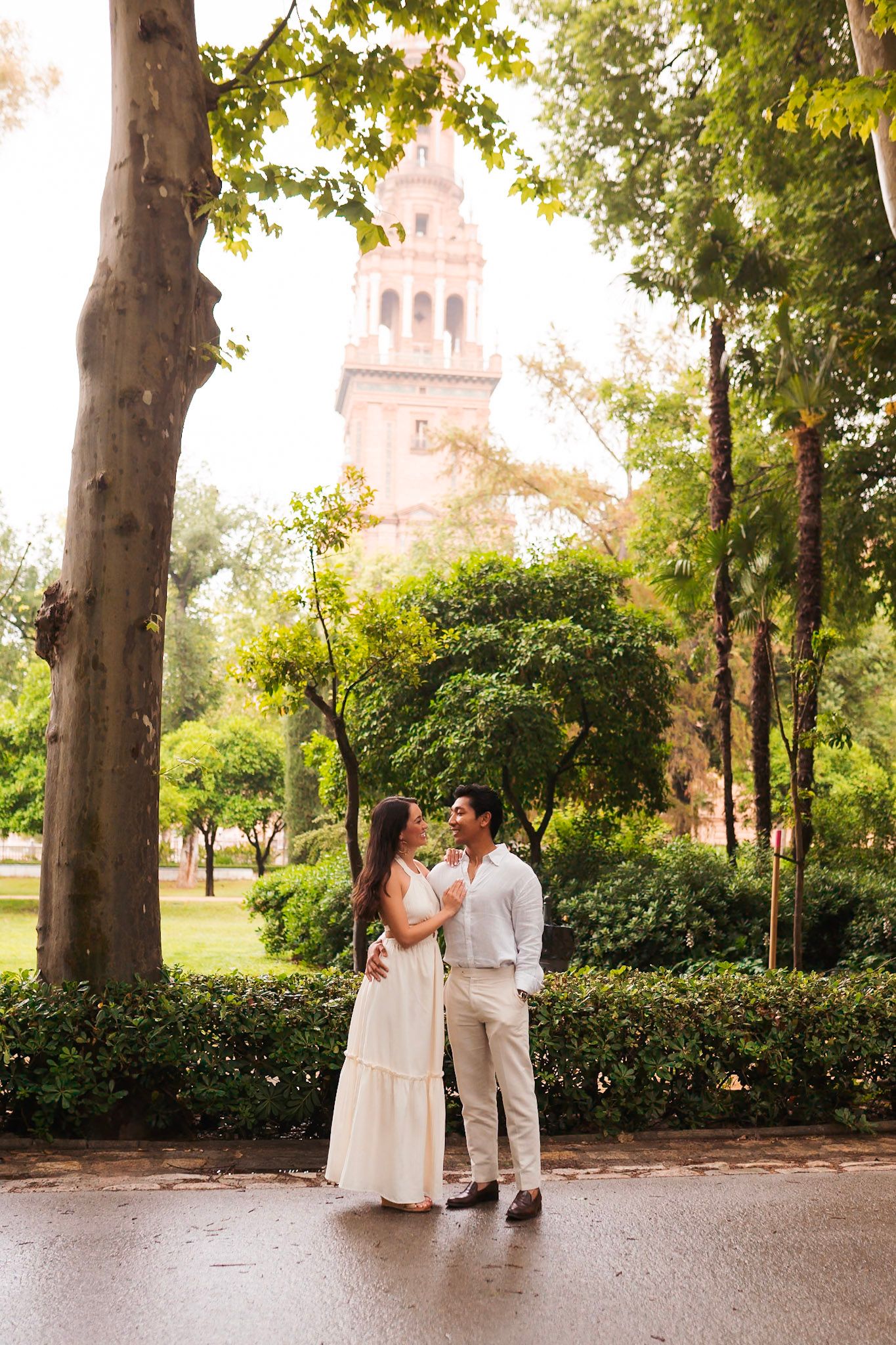 Romantic couple embracing in lush park with Plaza de España tower visible through trees, Seville.