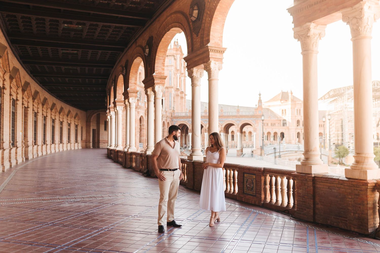 Couple in white dress and beige pants share a tender moment in sunlit arched colonnade at Plaza de España, Seville.